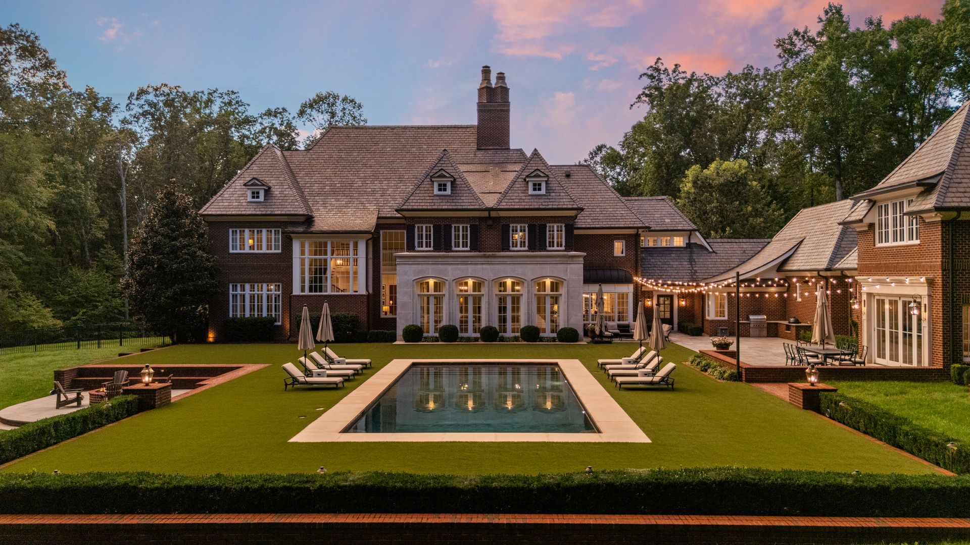 Evening view of a large brick house with a tiled roof, surrounded by trees, featuring a rectangular pool in a green lawn, lounge chairs, umbrellas, string lights, and outdoor seating areas.