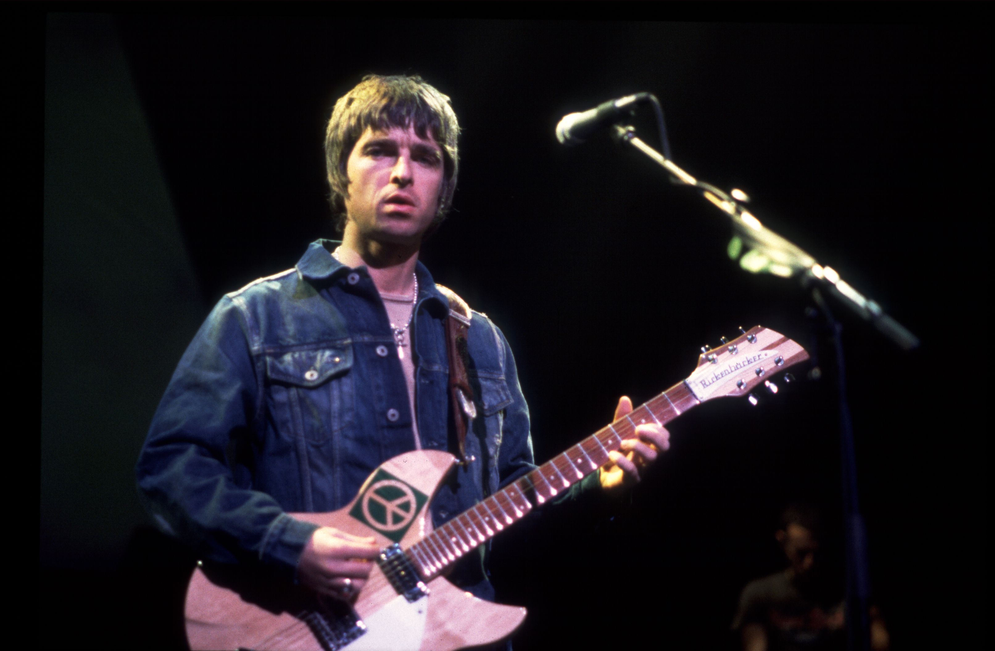 Man wearing a dark denim jacket playing a Rickenbacker guitar with a peace symbol on the body, standing in front of a microphone on a dark stage.