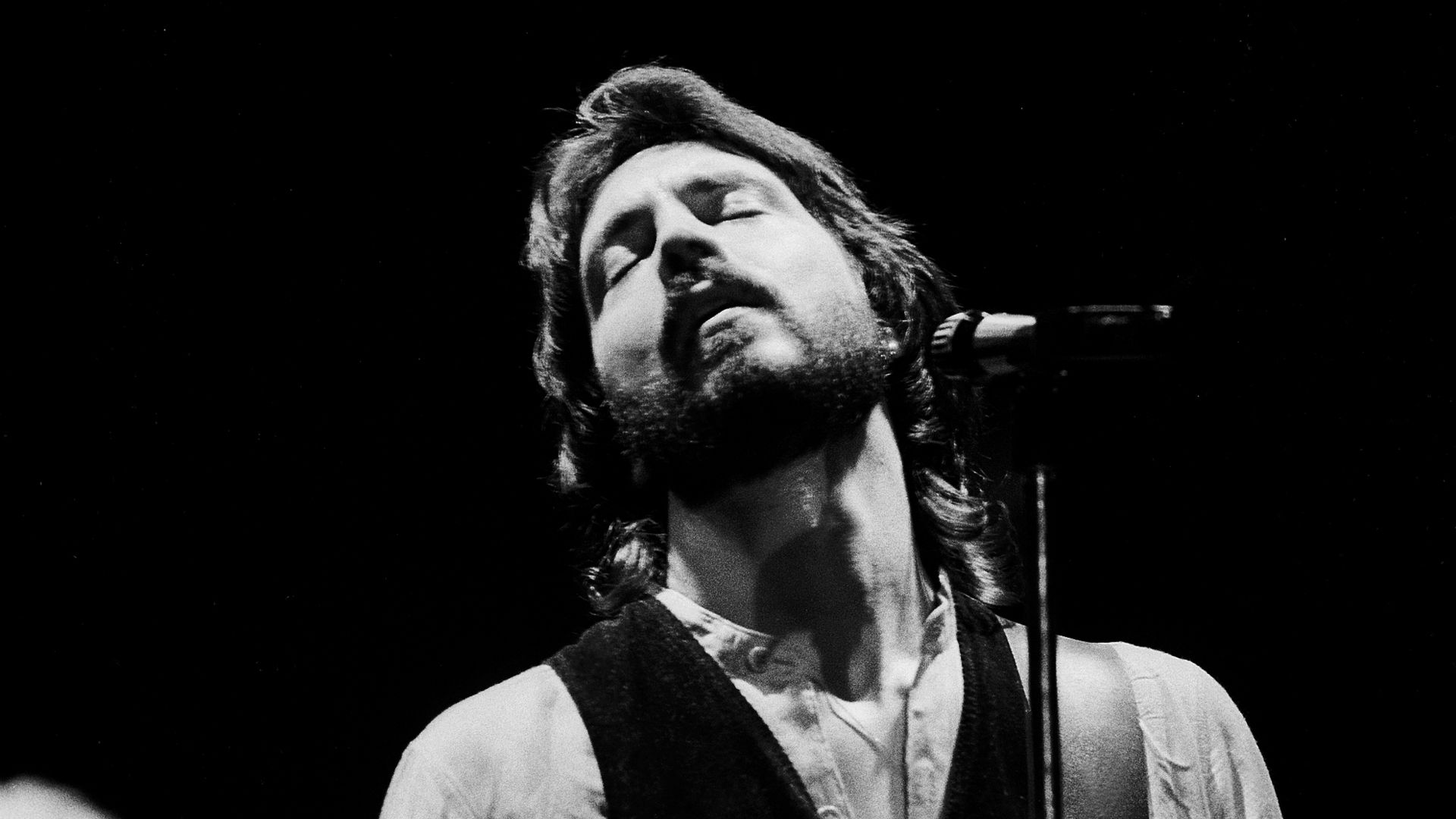 Black-and-white photo of a bearded male musician on stage, eyes closed, singing into a microphone while playing a light-colored electric guitar; wearing a vest over a light shirt.