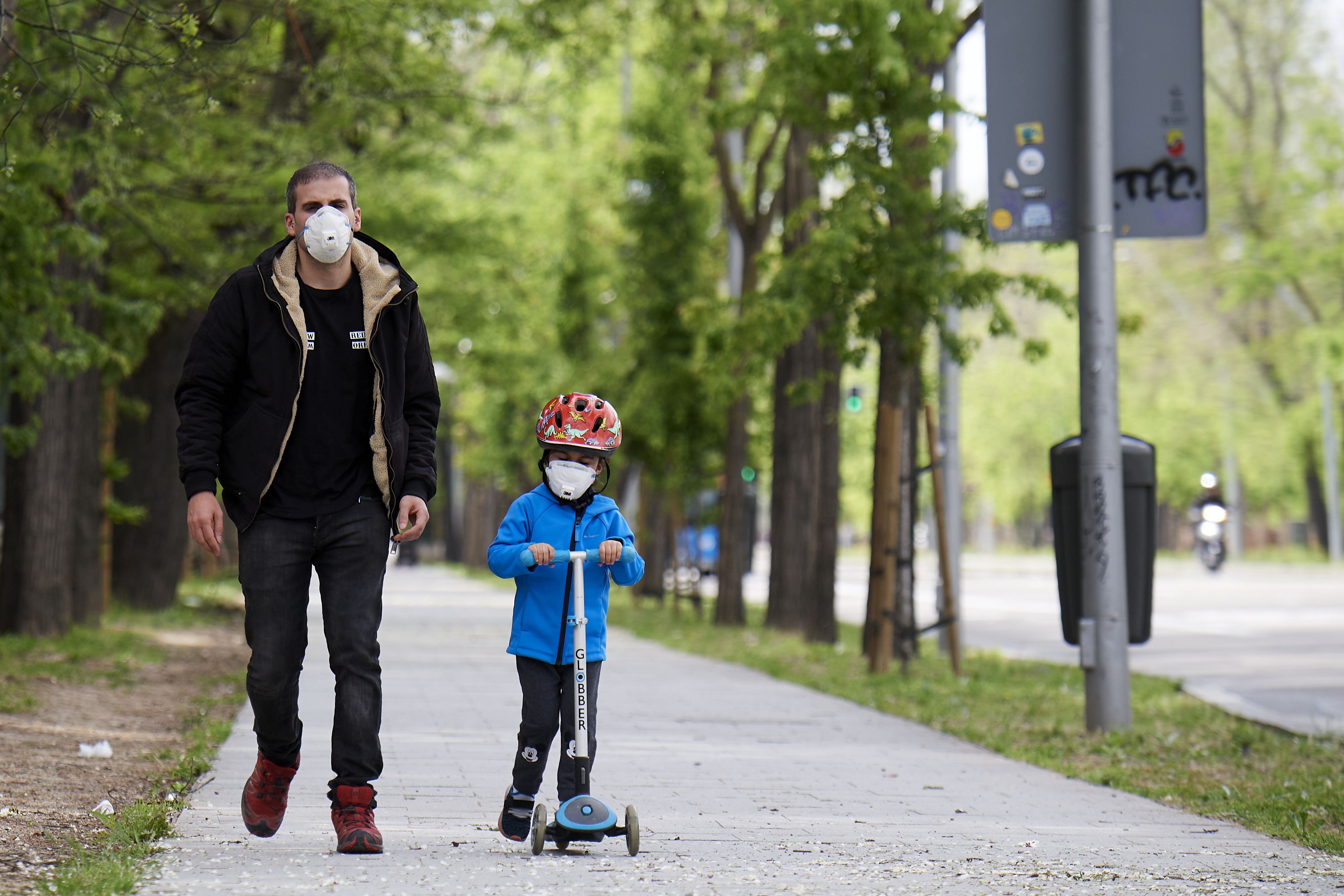In this image, a boy wears a mask on his scooter while walking with a masked man 