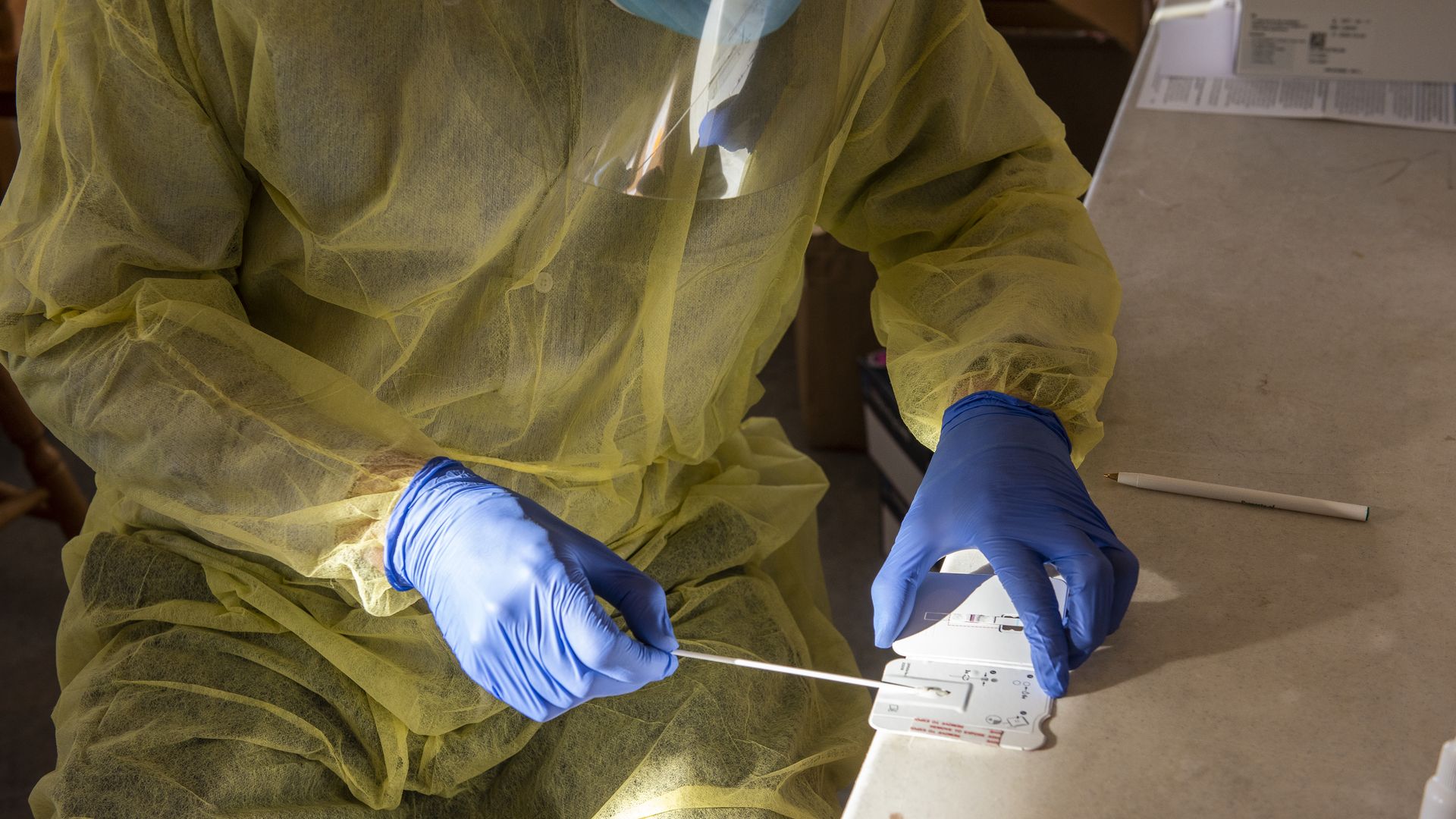 A health care worker wearing gloves and PPE places a swab in a COVID-test that rests on the edge of a table