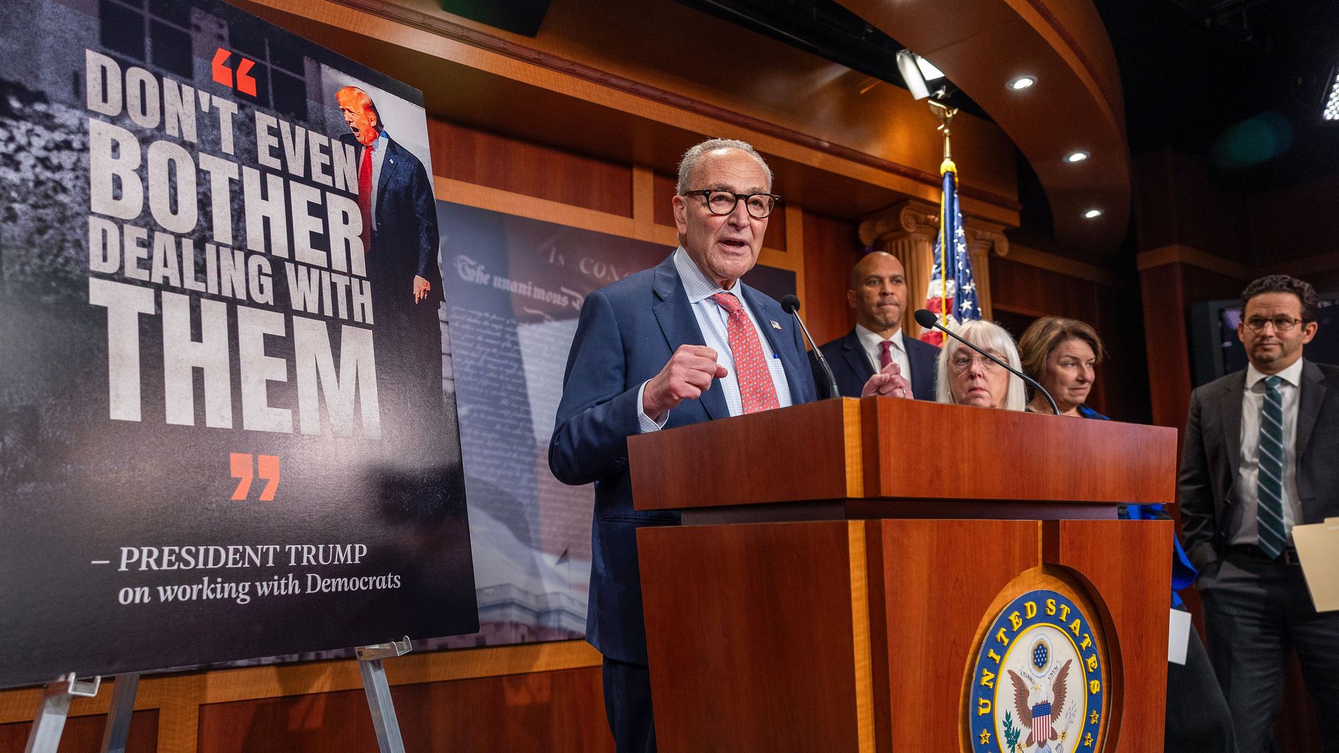 Chuck Schumer — wearing a blue suit, a red tie and glasses — speaks from behind a podium next to a large sign with a photo of Trump and text that says "'DON'T EVEN BOTHER DEALING WITH THEM' - PRESIDENT TRUMP on working with Democrats."