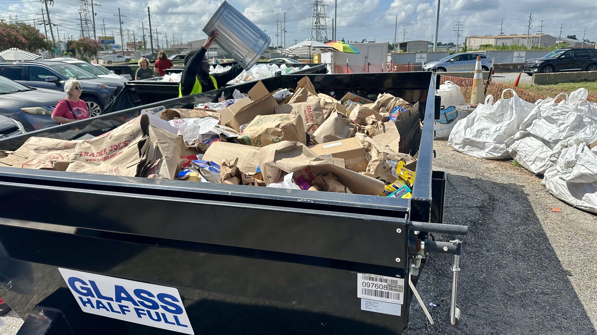 Worker in a neon safety vest tosses a metal lid into a large black dumpster overflowing with crumpled paper bags, cardboard, and trash; a woman in a pink shirt watches in a parking lot.