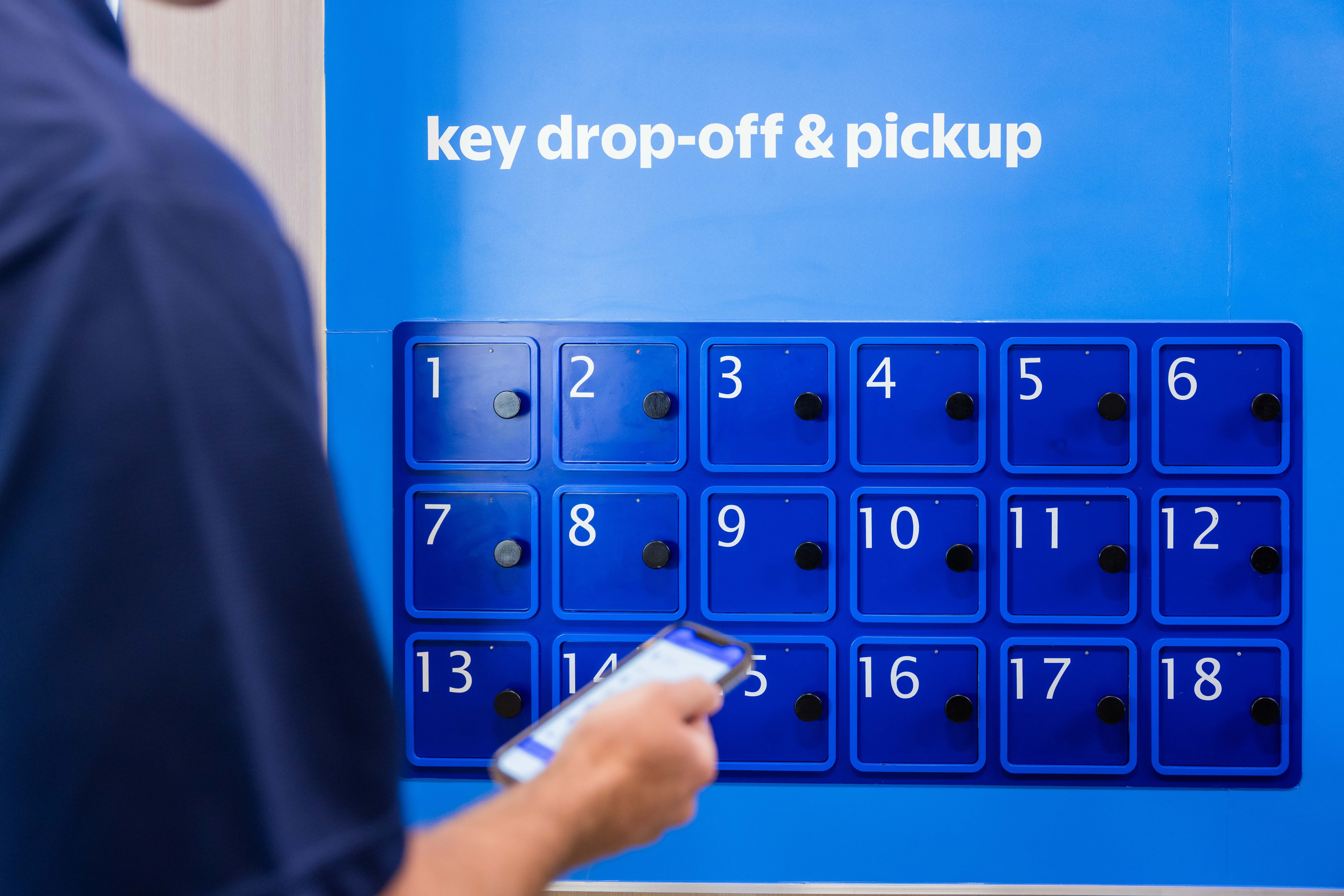 Blue key drop-off and pickup lockers numbered 1 to 18 on a blue background with a person holding a smartphone in the foreground.