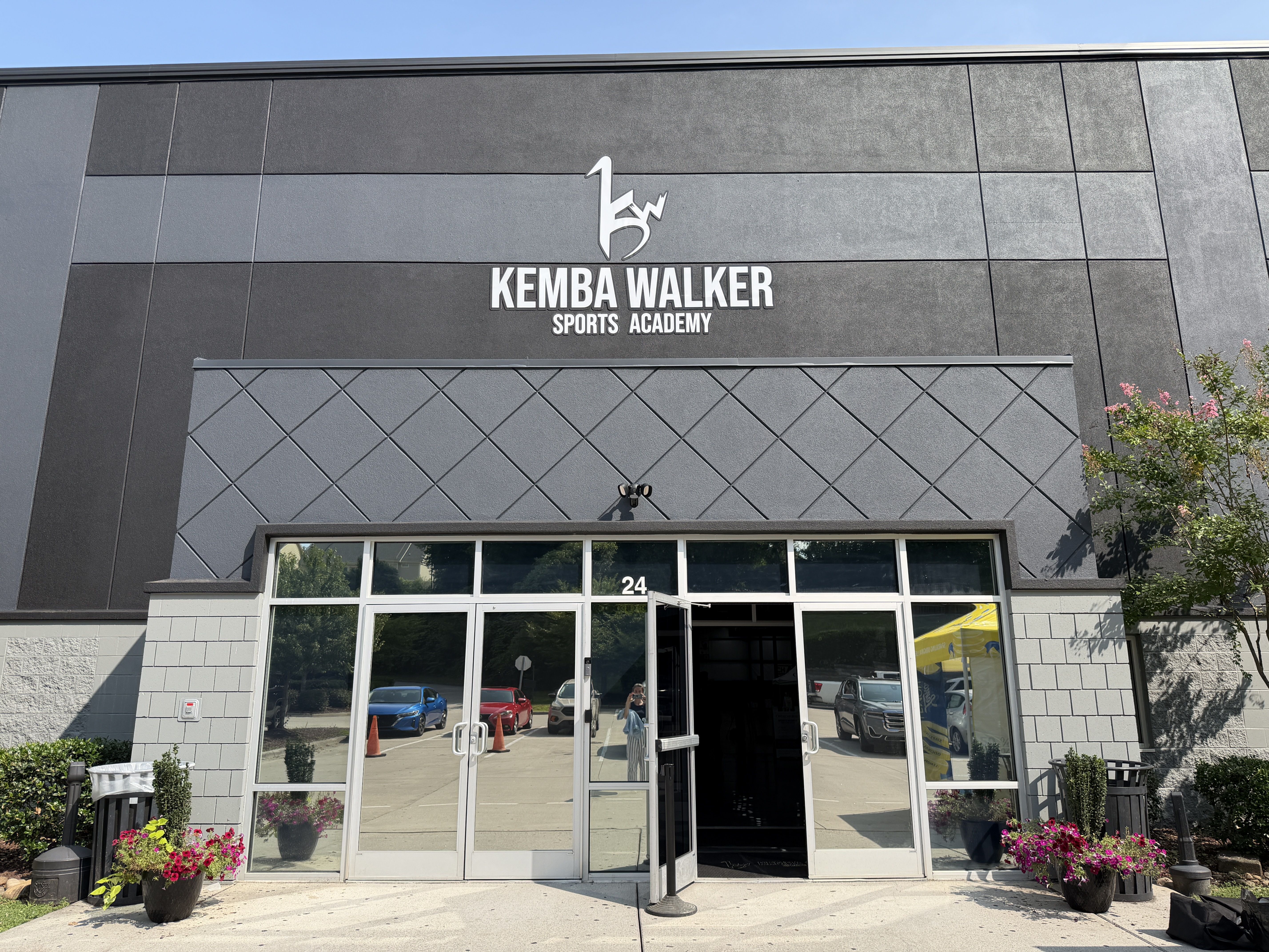 Exterior of a gray, modern sports academy building with a large white sign reading "KEMBA WALKER" and "SPORTS ACADEMY" above a stylized logo. Glass doors, parked cars, blue sky, and potted flowers.