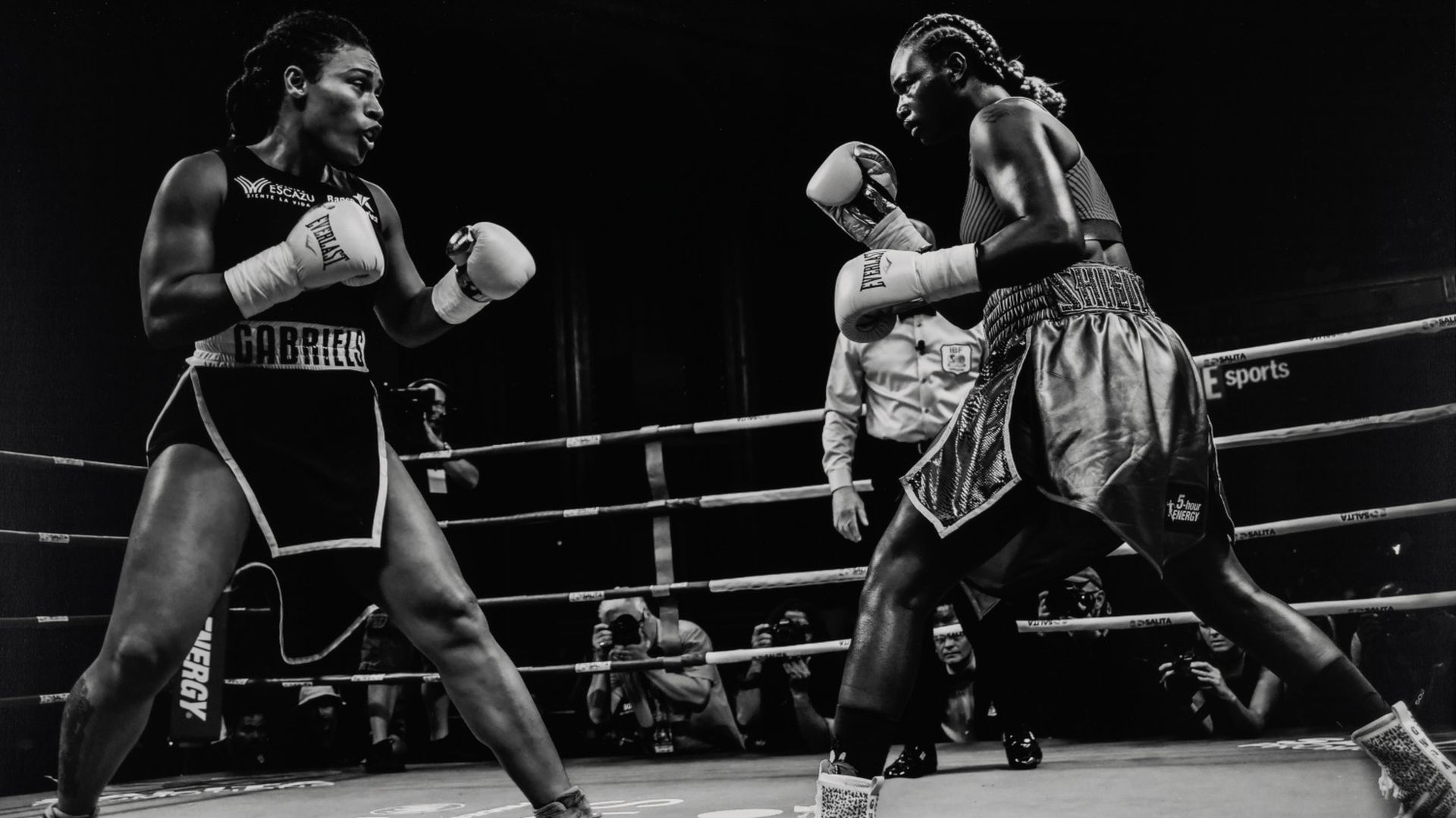 Black-and-white photo of two female boxers in a ring with gloves up, facing each other intensely, with a referee and photographers visible in the background.