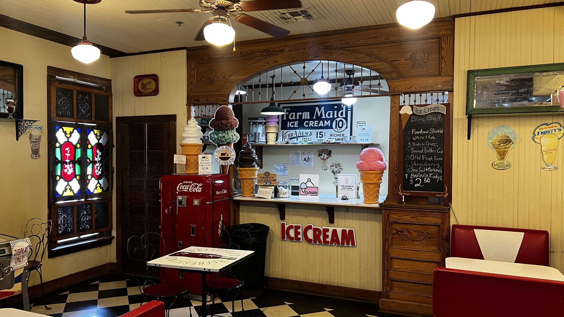 Vintage ice cream shop interior with yellow paneled walls, checkered black-and-white floor, wooden arch, ceiling fans, globe pendant lights, and oversized ice cream cone displays.