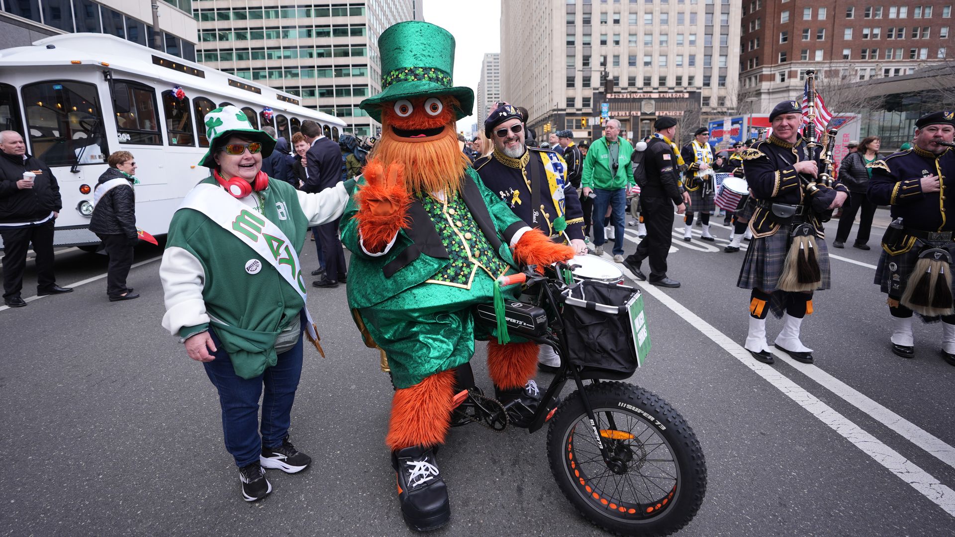 Urban parade with tall office buildings. A bright orange leprechaun on a green suit rides a small bike, waving. Nearby a person in a shamrock hat smiles as bagpipers in kilts march.