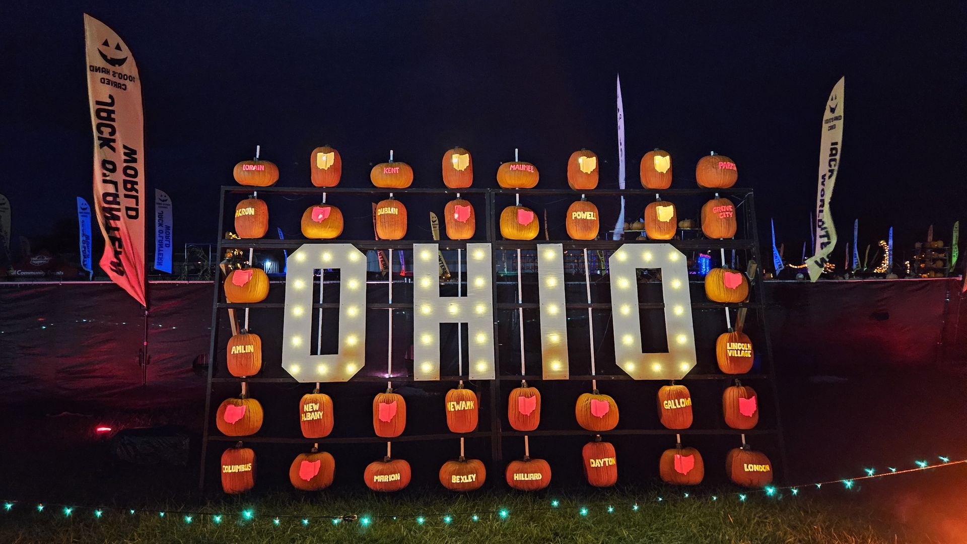 A sign reading "Ohio" lit up, with fake pumpkins on stakes surrounding it, featuring carvings of city names and the state's outlines