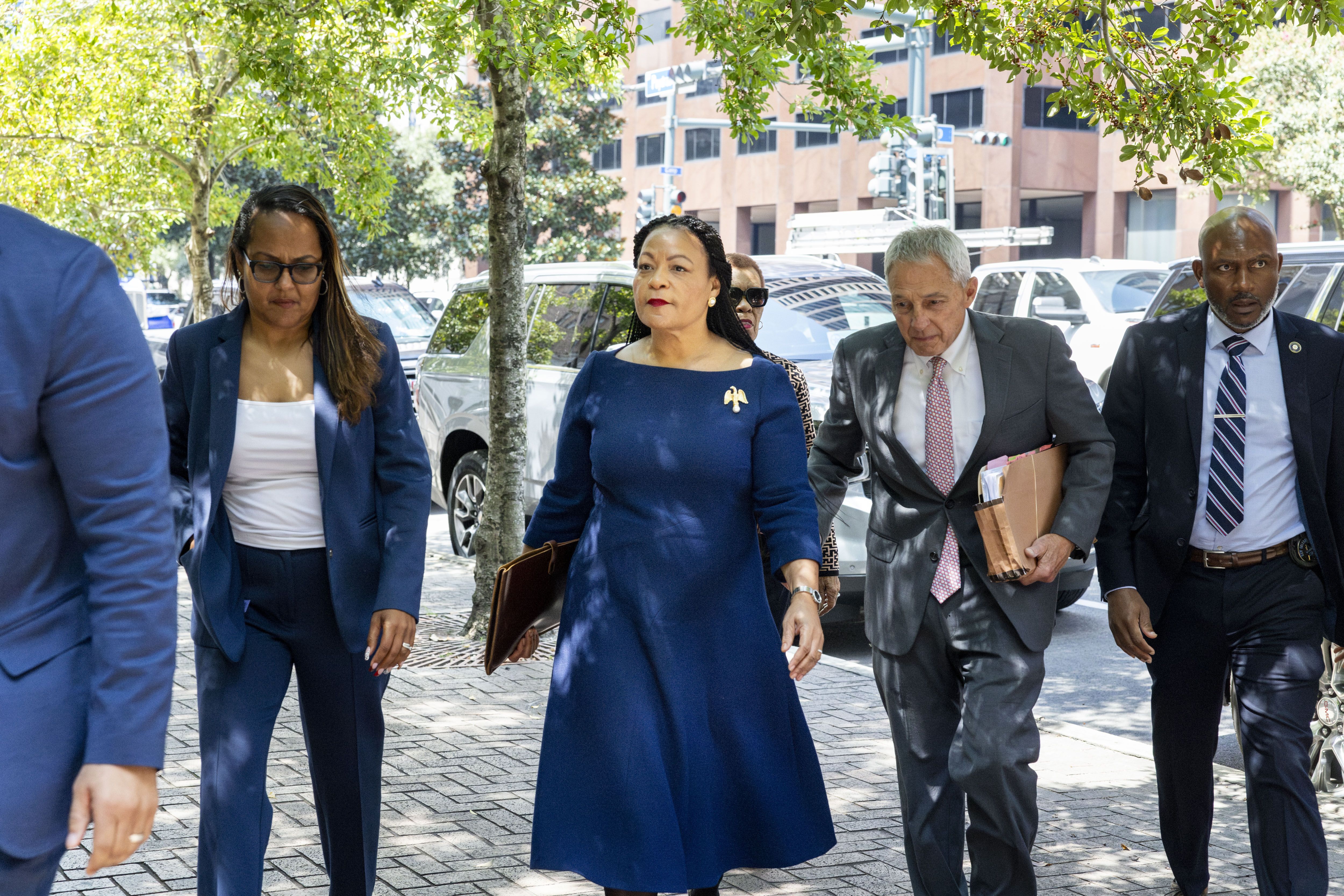 A group of professionals, including a woman in a blue dress and a man in a gray suit holding folders, walk on a city sidewalk under leafy green trees near parked cars.