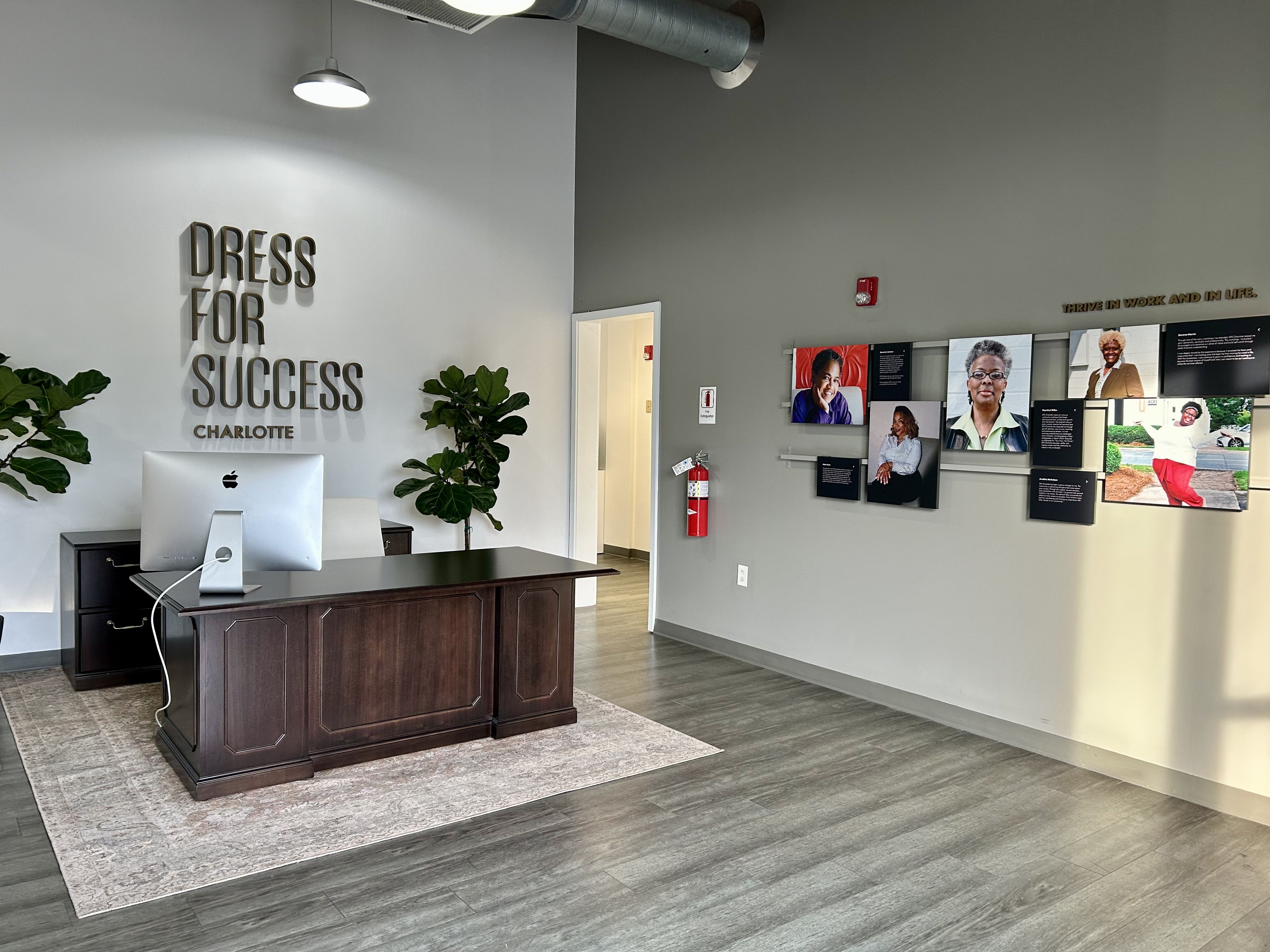 Office reception with dark wooden desk, Apple computer, two green plants, and wall text "DRESS FOR SUCCESS CHARLOTTE." Portraits and text displays on adjacent gray wall.