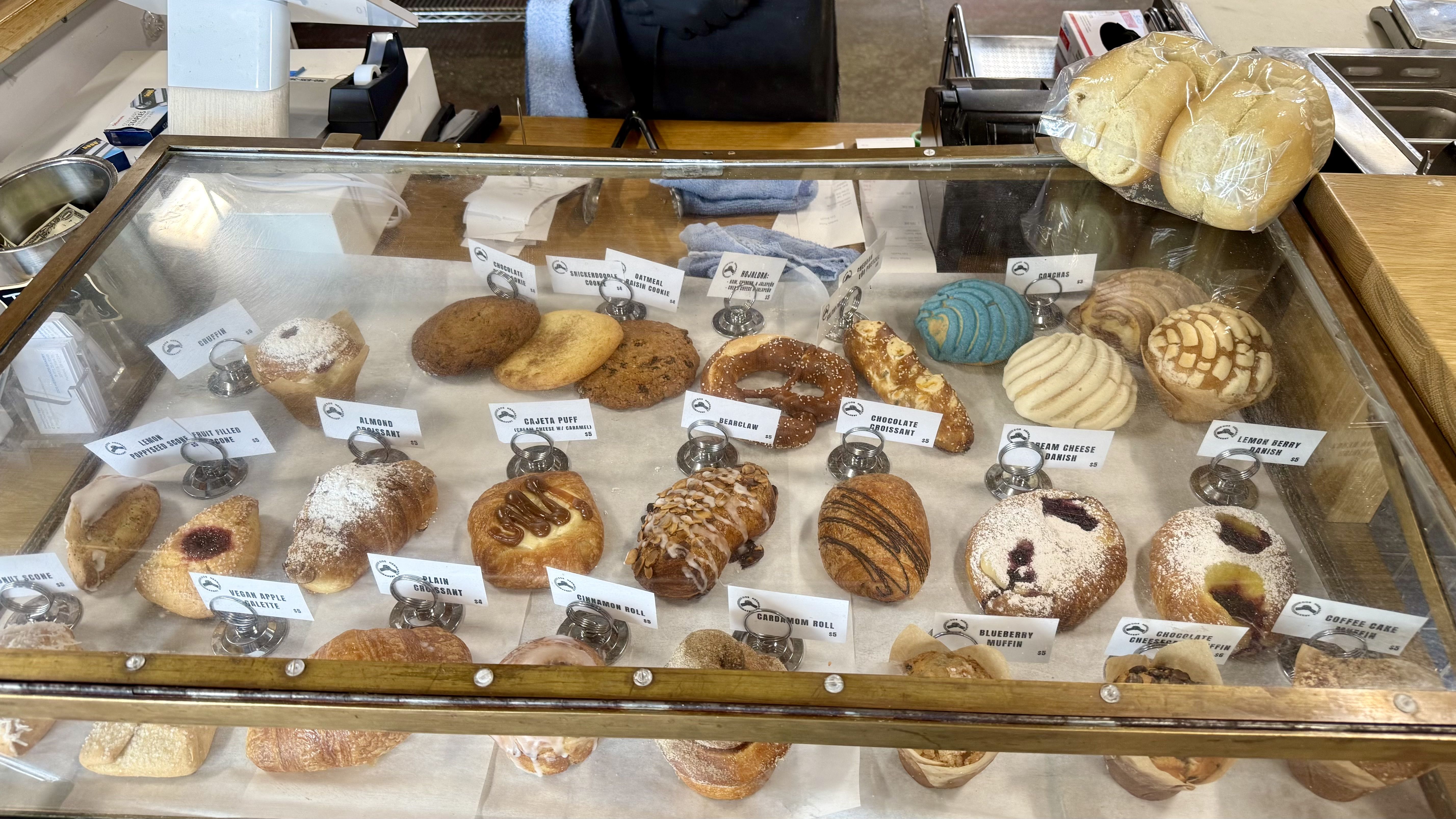 Glass pastry case filled with labeled baked goods including croissants, muffins, cookies, and danishes inside Dos Hermanos Bakery.