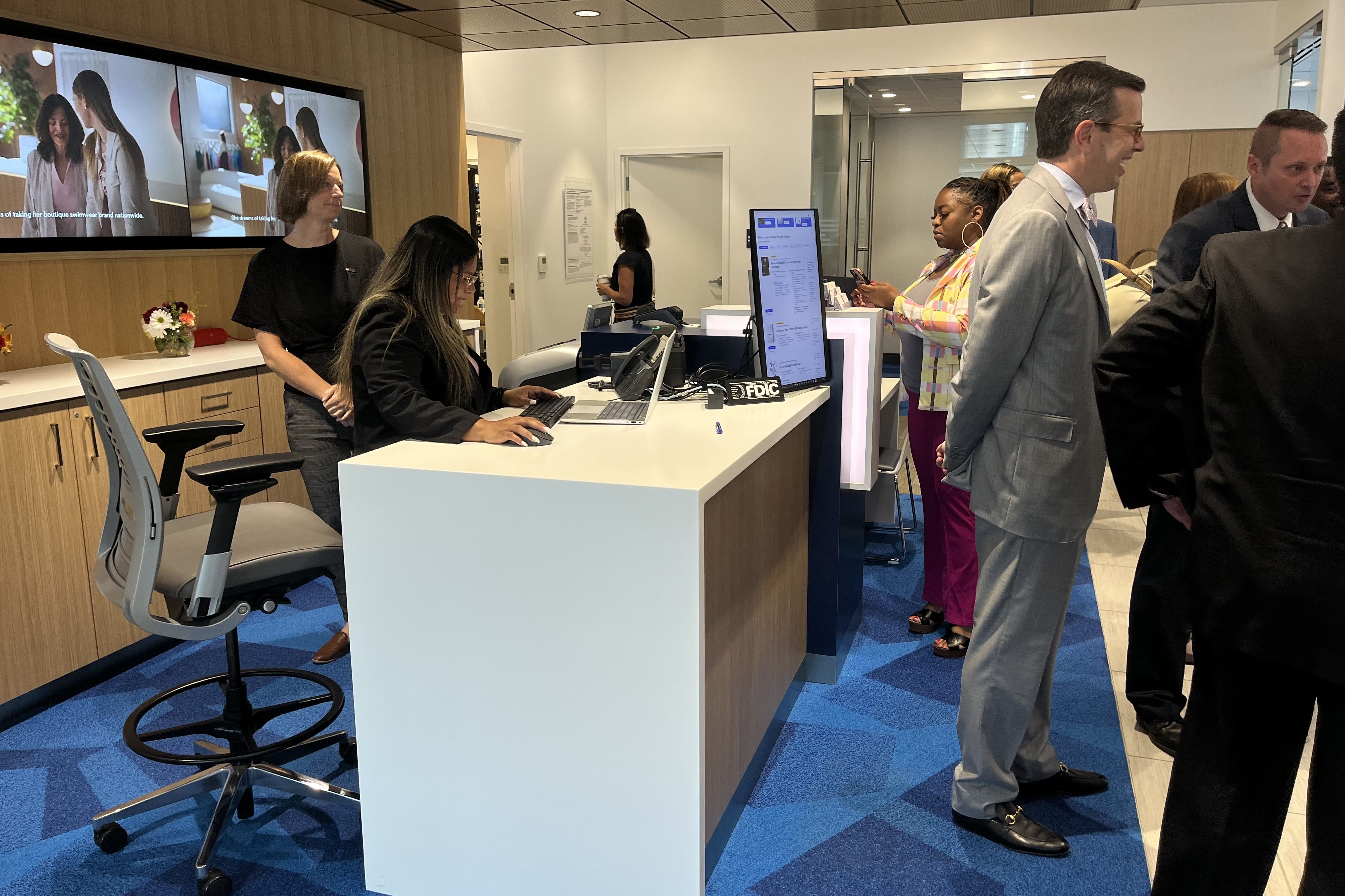 Bank lobby with multiple people: a woman working on a laptop behind a white desk, a woman standing behind her, a man in a gray suit talking, and others waiting, blue carpet and wooden accents.
