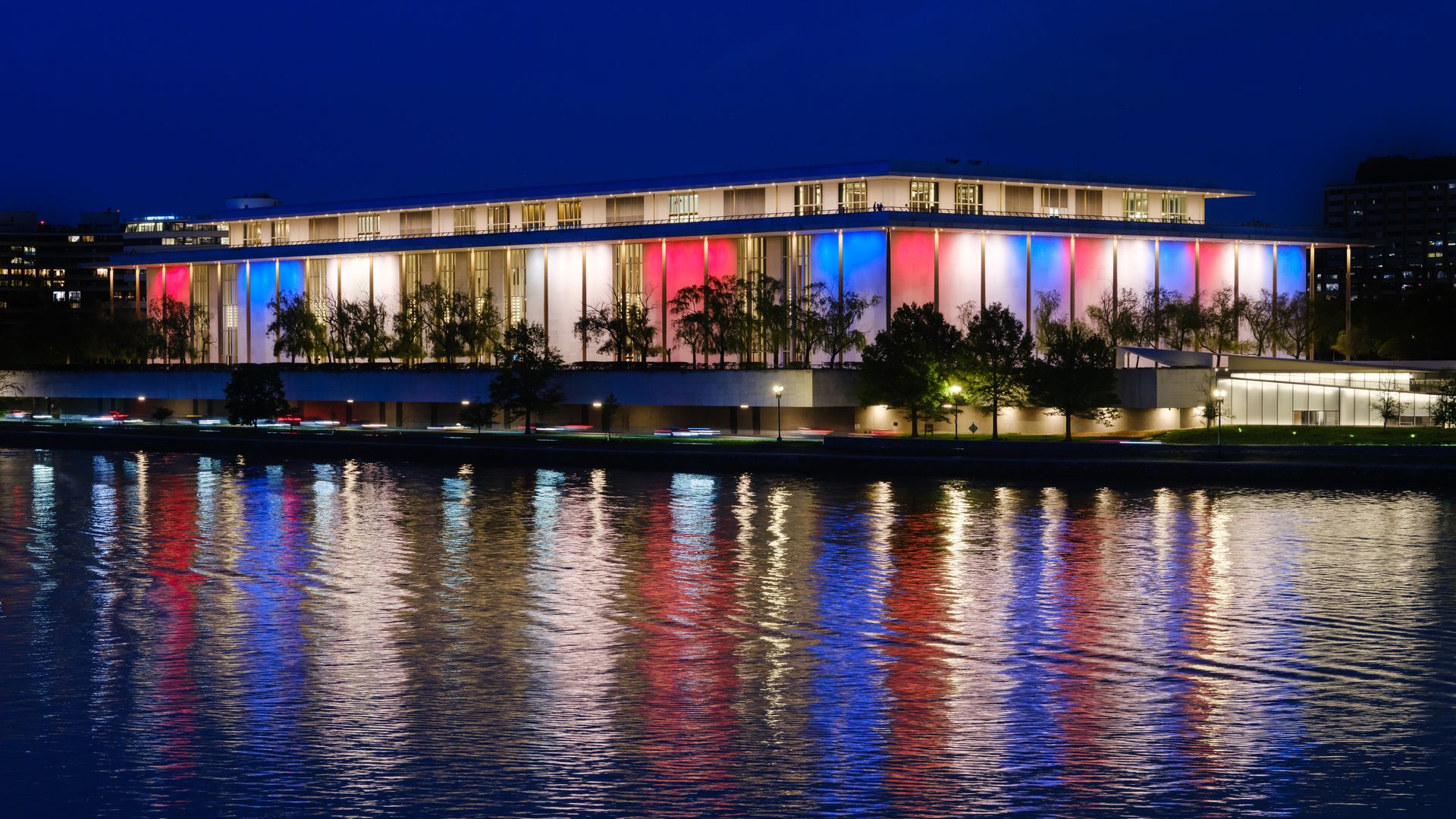 The John F. Kennedy Center for the Performing Arts is lit in red, white, and blue and reflected in the Potomac River at dusk in 2025.
