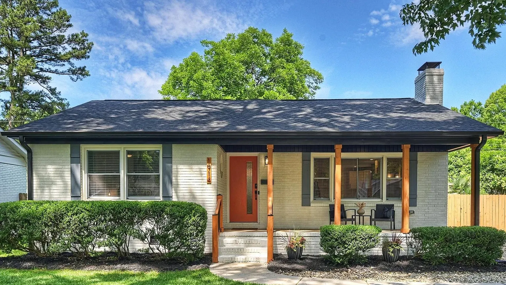 hot homes june 5 2026. Light brick ranch-style house with a dark roof, orange front door, and two wooden porch columns. Front shrubs, a small seating area, and a green lawn under a blue sky.