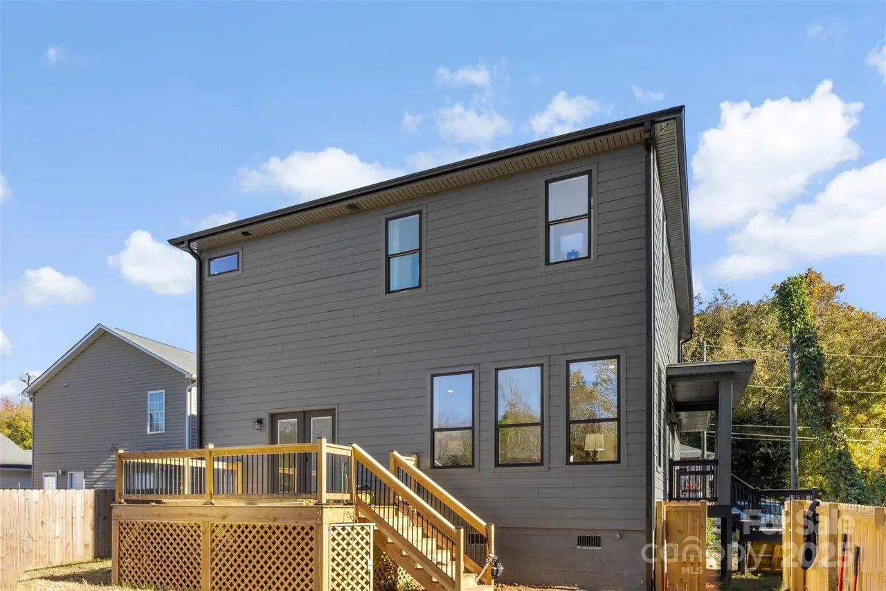 Two-story gray house with six windows, wooden deck, and stairs with railing under a blue sky with white clouds, next to a wooden fence and trees in the background.