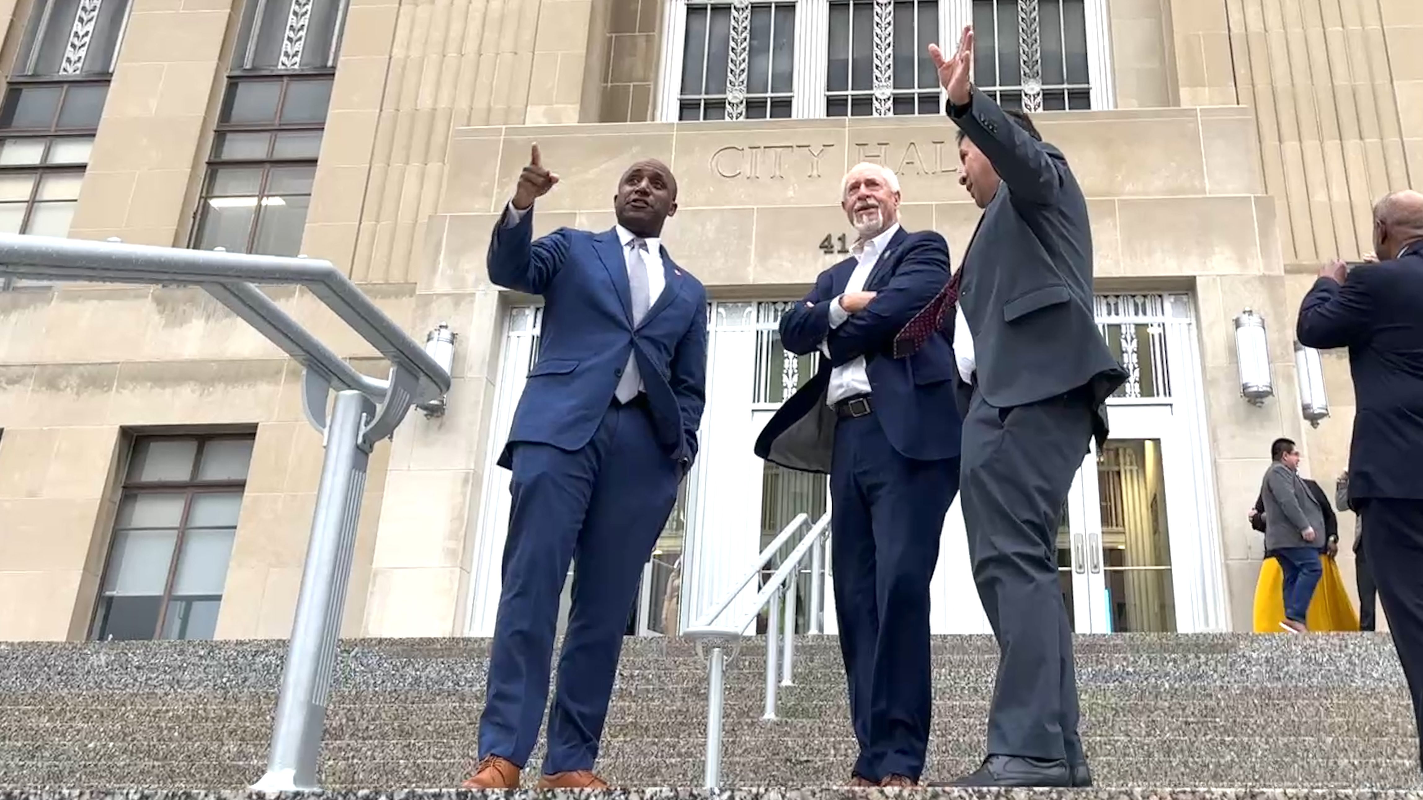 Quinton Lucas, Kevin O'Neill and Mario Vasquez in suits stand on the steps of city hall, one pointing upward, another with arms crossed, a third gesturing with raised hand. The facade reads CITY HALL behind them.