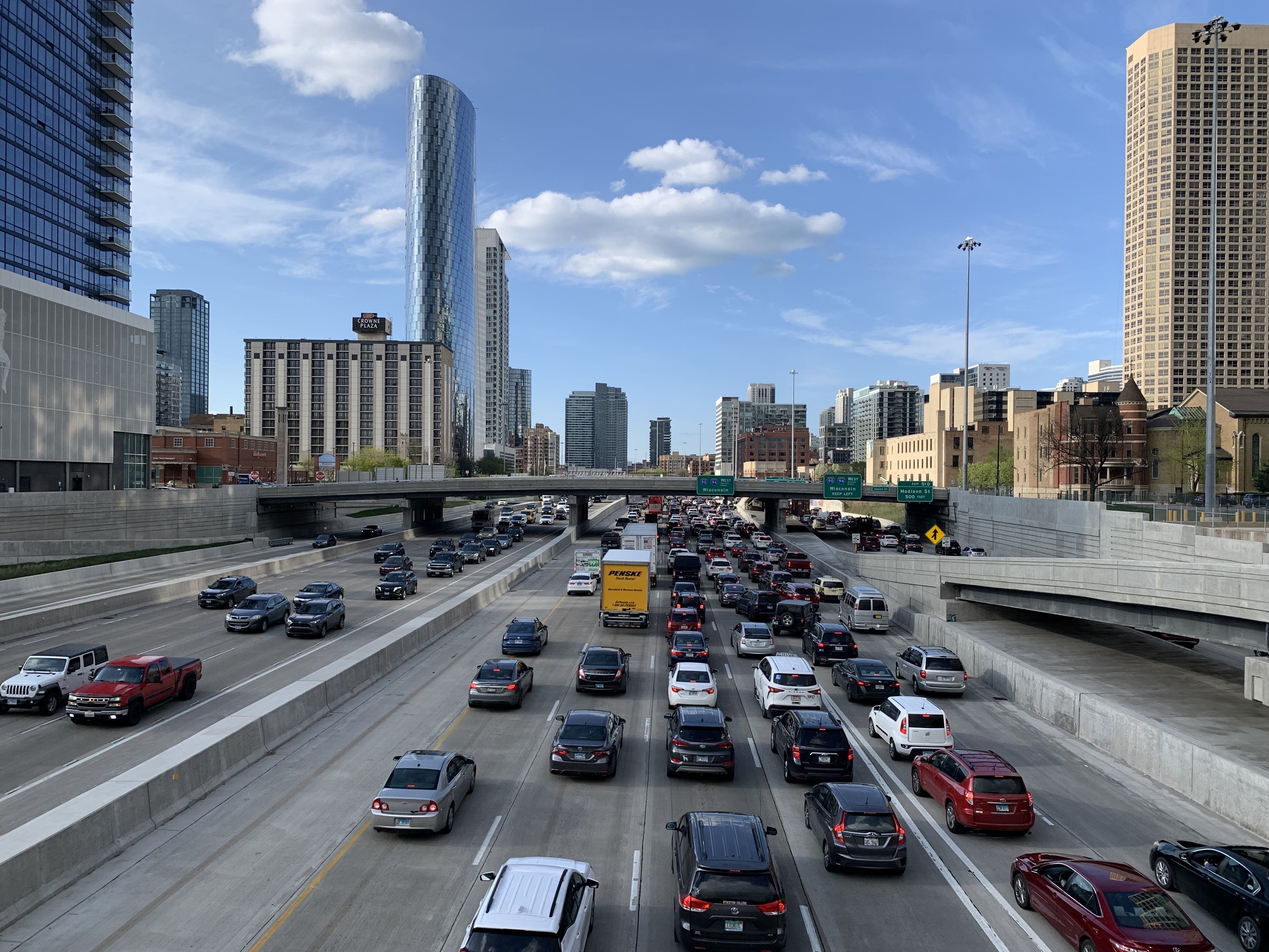 Busy multi-lane highway during daytime with heavy traffic in an urban area, skyscrapers including a shiny curved glass building, a blue sky, and some white clouds overhead.