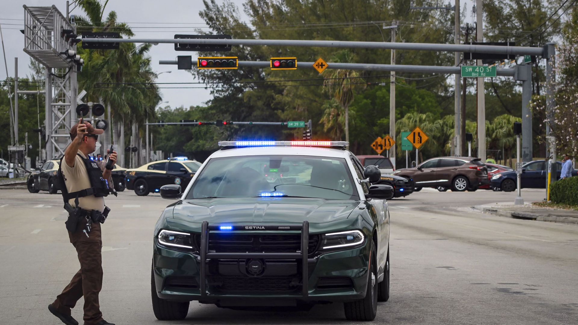 Police officer directing traffic near a green police car with flashing red and blue lights at an intersection with traffic signals and street signs showing SW 40th St and Bird Rd.