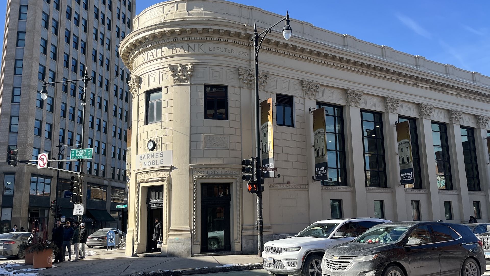 Beige historic building with "Noel State Bank 1920" inscription, now a Barnes & Noble bookstore, located on a sunny city corner with pedestrians and cars waiting at a crosswalk.