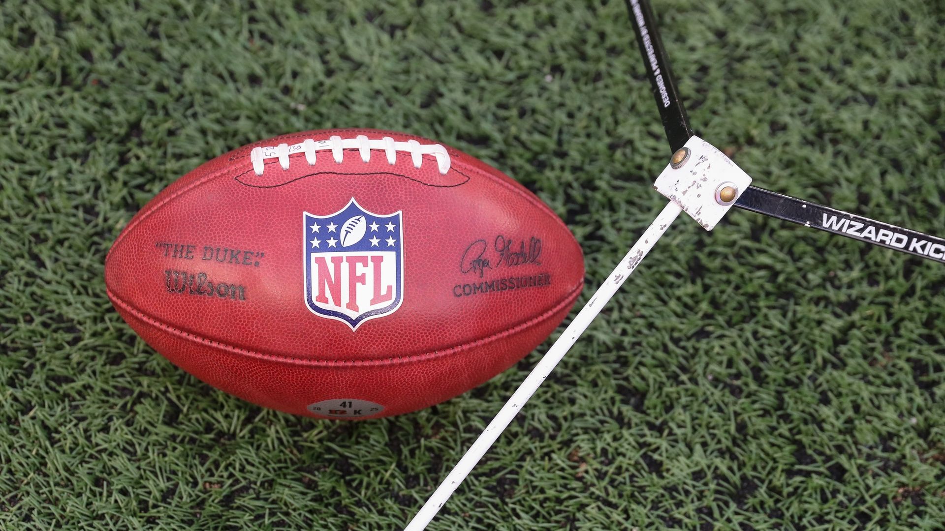 Red NFL football on green turf with white laces and the NFL shield, plus signatures. A white kicking trainer labeled "WIZARD KICK" intersects near the ball.