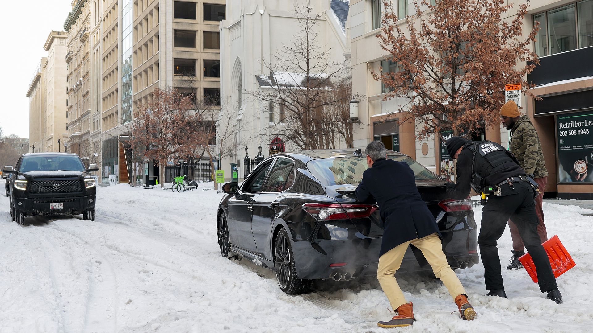 Two people push a stuck car in a snowed-in downtown street