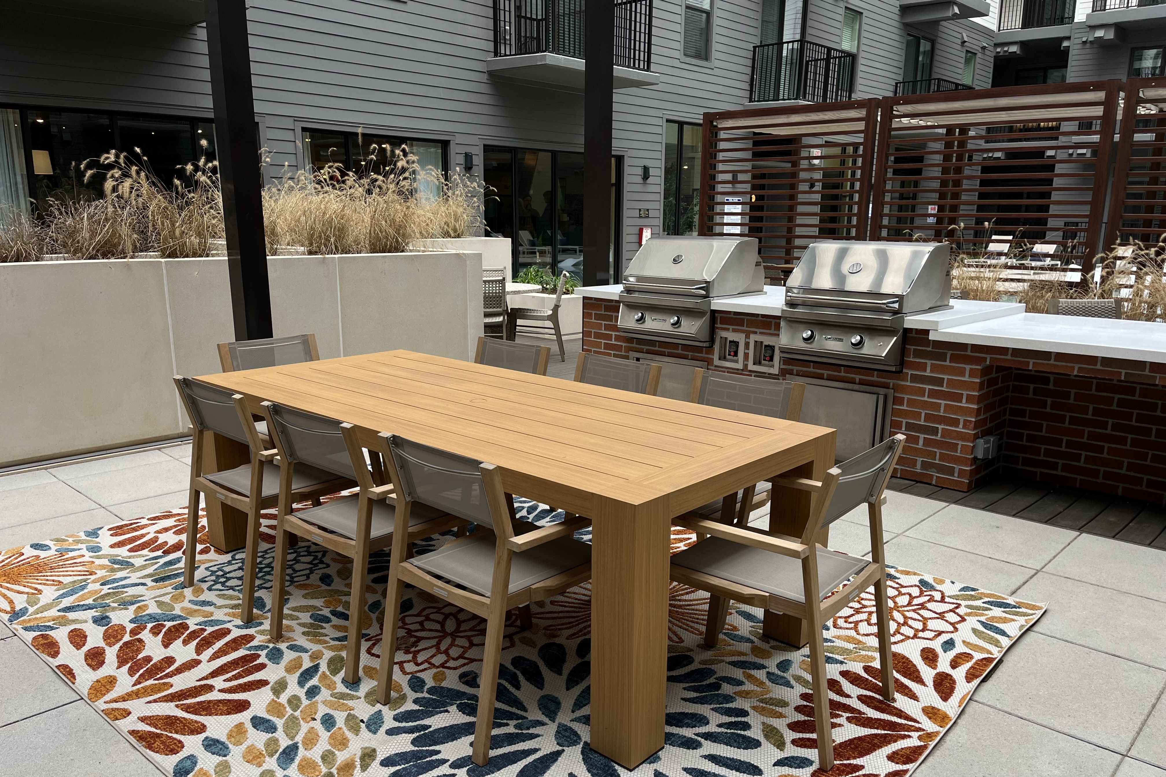 Outdoor dining area with a wooden table surrounded by eight gray chairs on a colorful floral rug, two stainless steel grills against a brick counter, and gray apartment building walls in the background.