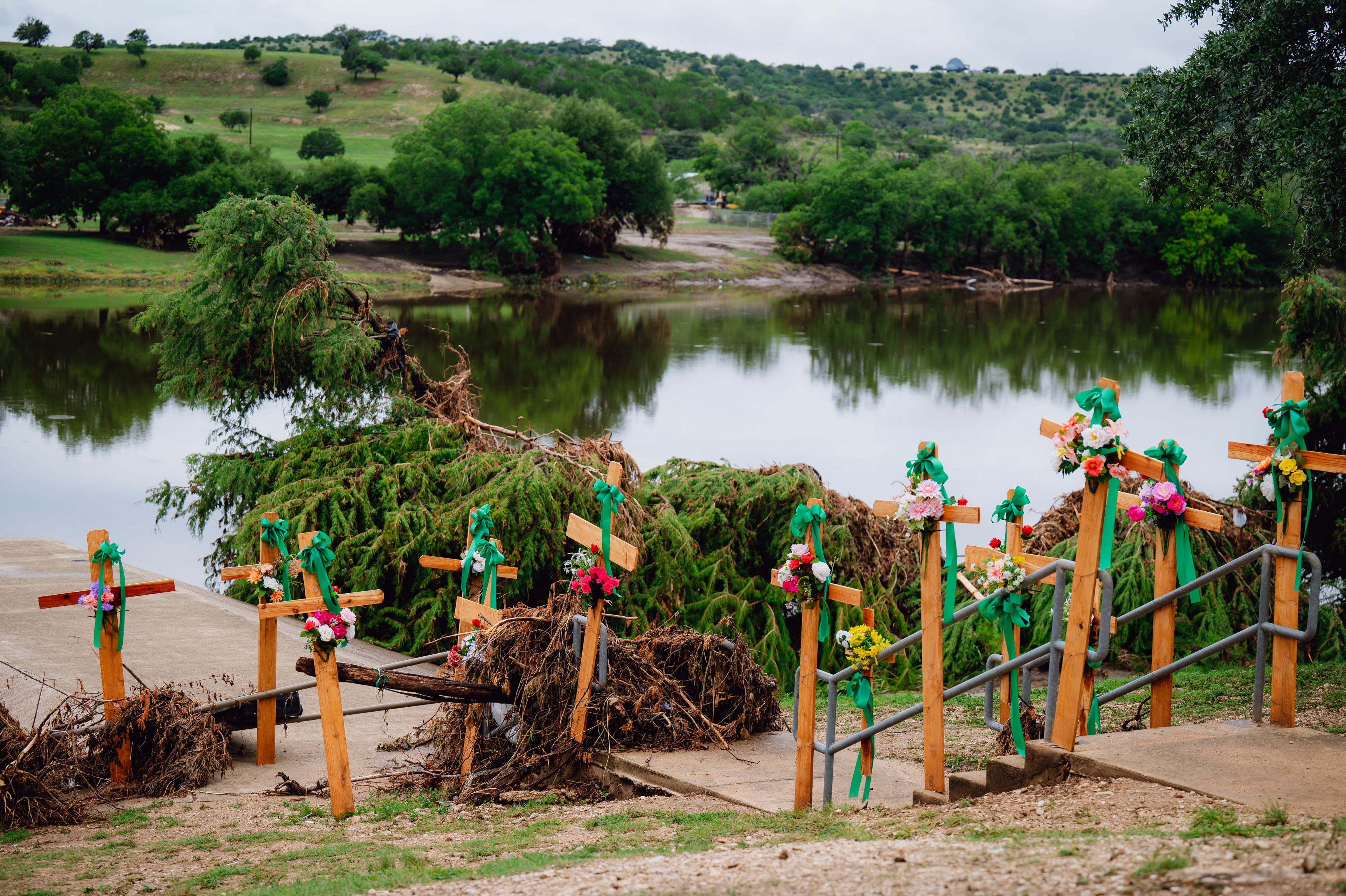 Crosses line the Guadalupe River at Guadalupe Park in Kerrville in July 2025. The crosses are seen in front of mangled tree debris and with green ribbons tied around them.