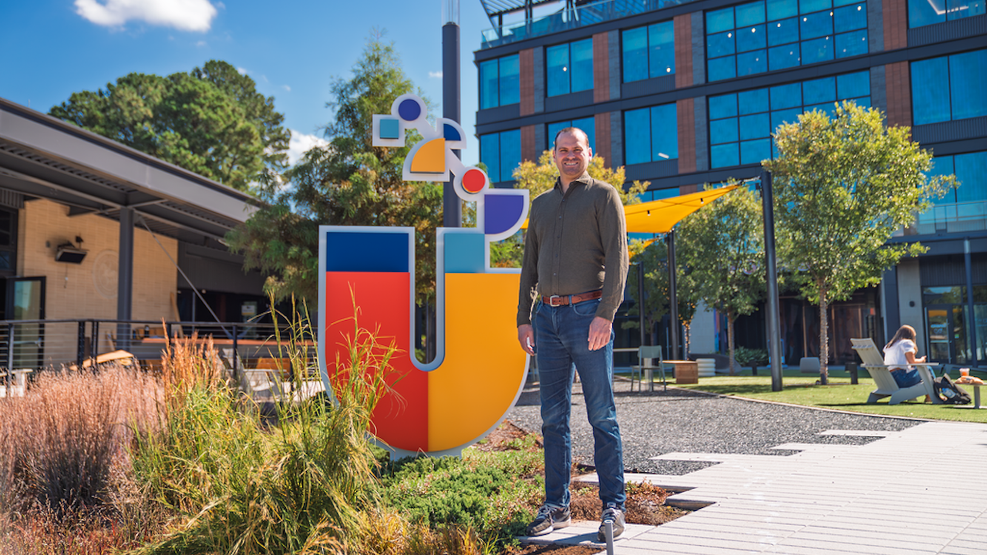 Man stands next to a large, colorful sculpture in a sunny modern campus courtyard; blue, red, yellow panels with a glass-fronted building and trees in the background.