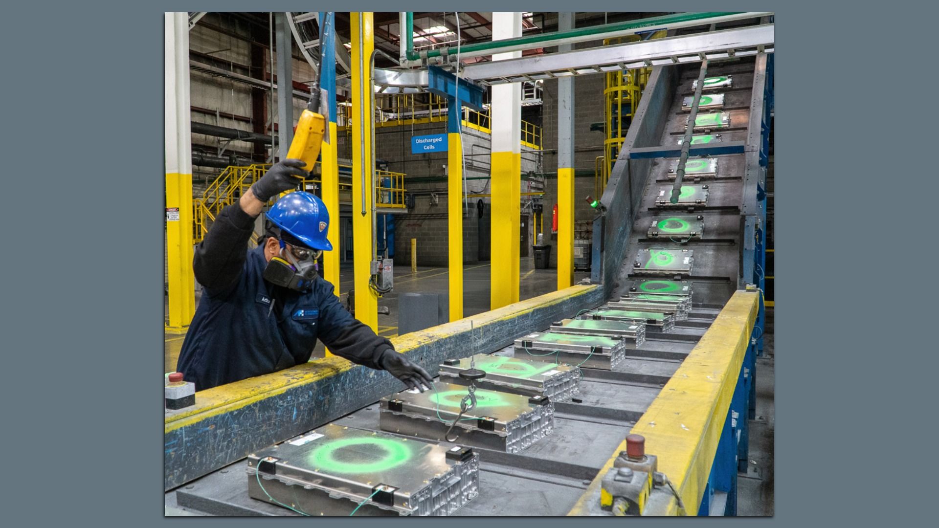 Industrial factory: worker with blue hard hat and mask raises a yellow tool over a conveyor with metal trays bearing green circles; yellow safety rails and large machinery surround.