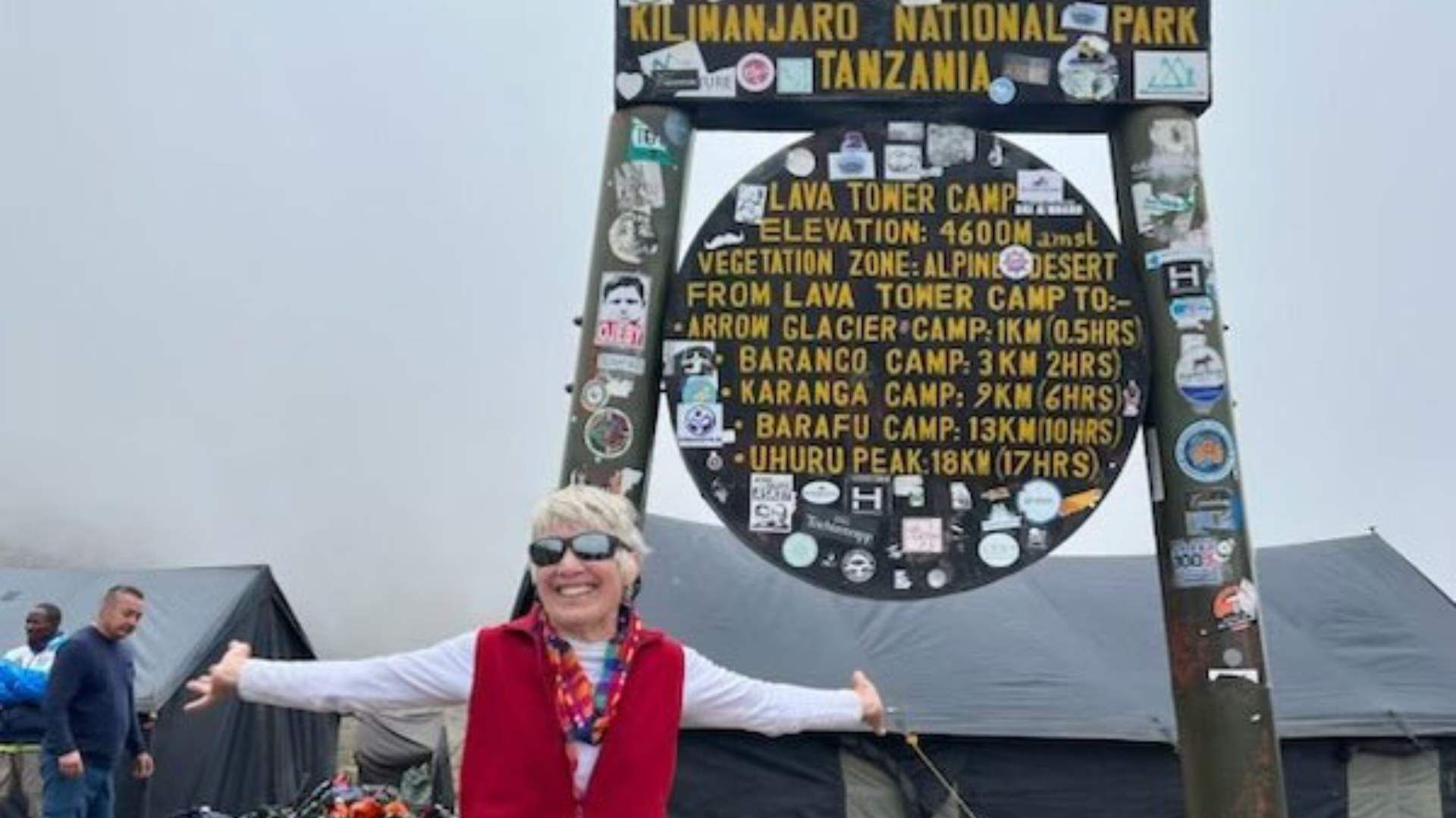 A smiling woman celebrates at Lava Tower Camp on Mount Kilimanjaro, surrounded by informational signage marking elevation and trails.