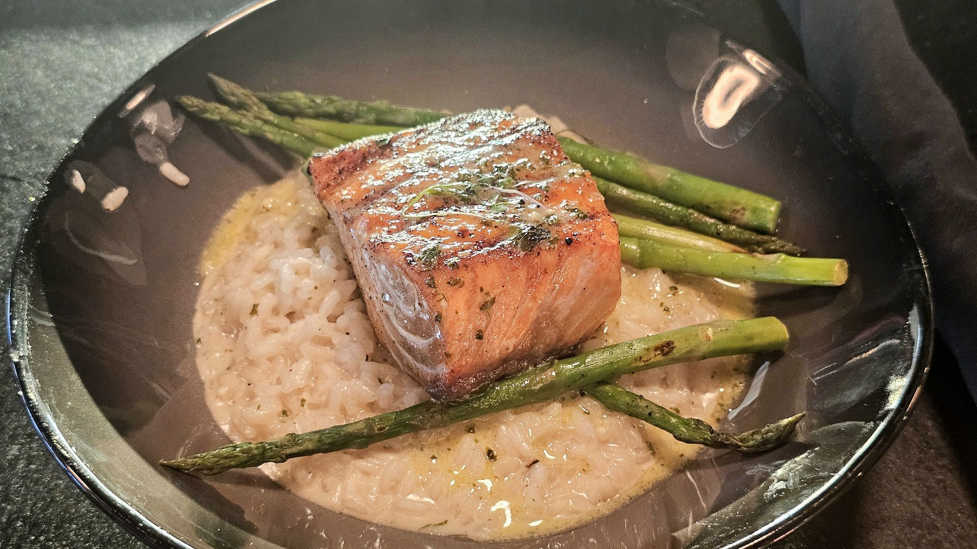 Pan-seared salmon fillet with herbs rests on creamy risotto, surrounded by steamed asparagus spears in a dark pot.