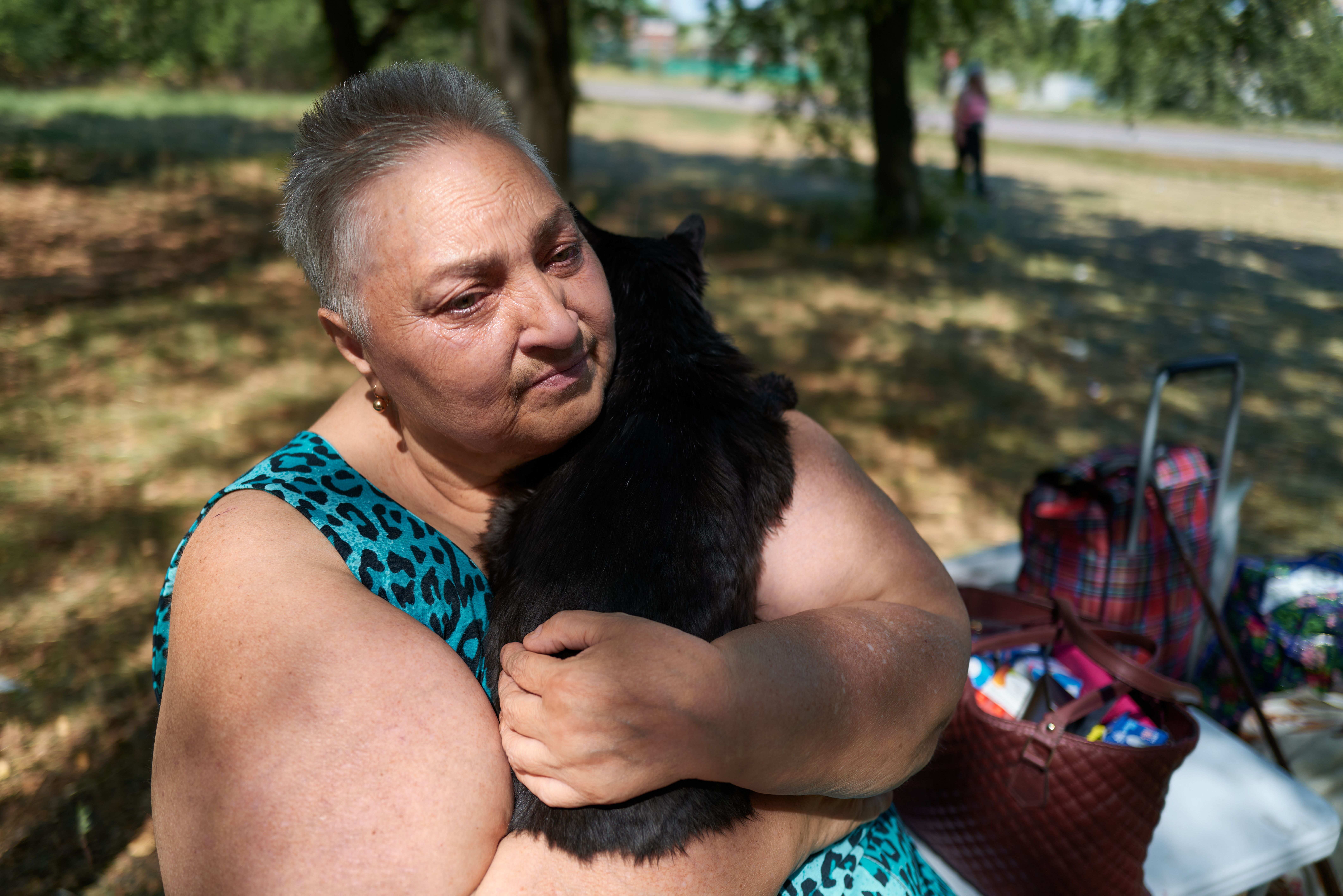 An elderly woman in a blue leopard print top holds and hugs a black cat outdoors in a shaded area with bags and a walker behind her.