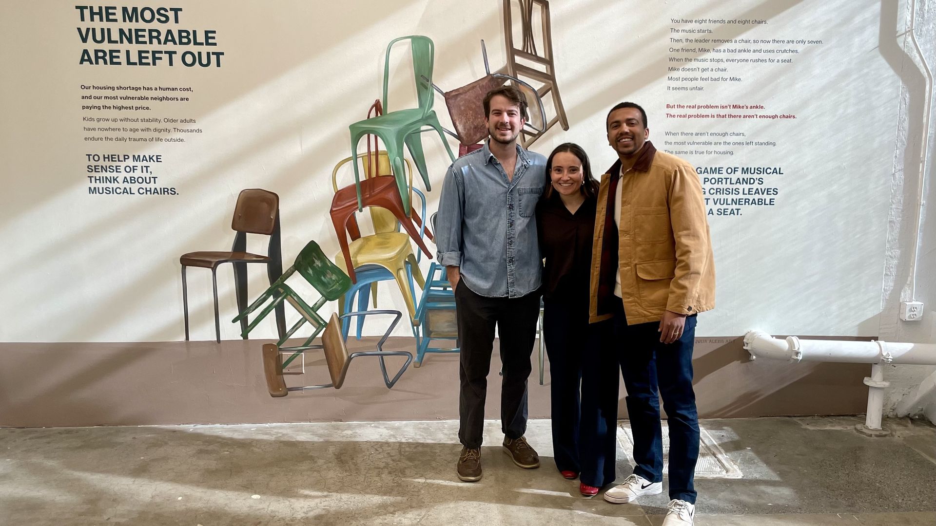 Three people smiling and standing in front of a mural of stacked colorful chairs and text about housing vulnerability and musical chairs illustrating housing crisis issues.