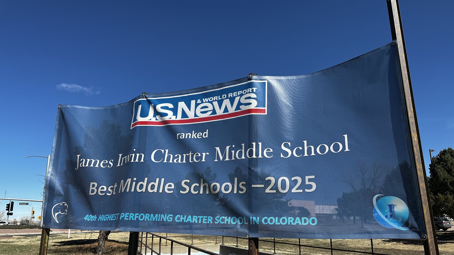 Blue outdoor banner with the "U.S. News" logo announcing James Irwin Charter Middle School ranked "Best Middle Schools – 2025" in Colorado; metal posts and a clear blue sky in the background.