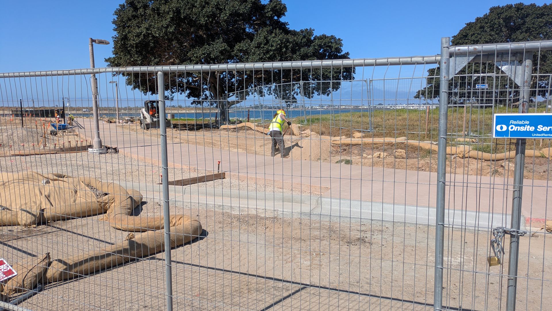 Construction site by the water with a metal chain-link fence, sandbags, and a worker in a yellow safety vest on a paved path; a large tree and blue sky with sea beyond.