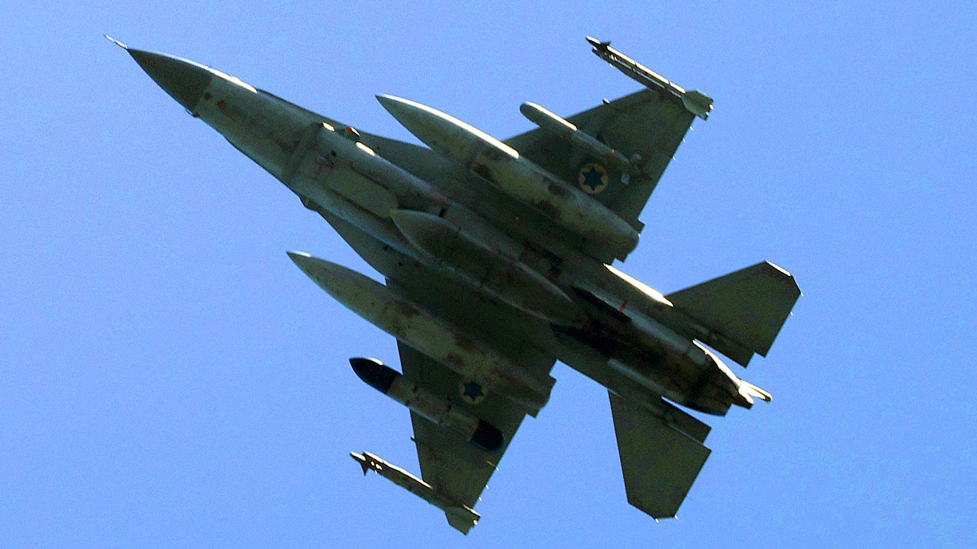 An Israeli Air Force F-16 flies above the buildings and hills of Yokneam Illit in northern Israel under a clear sky.