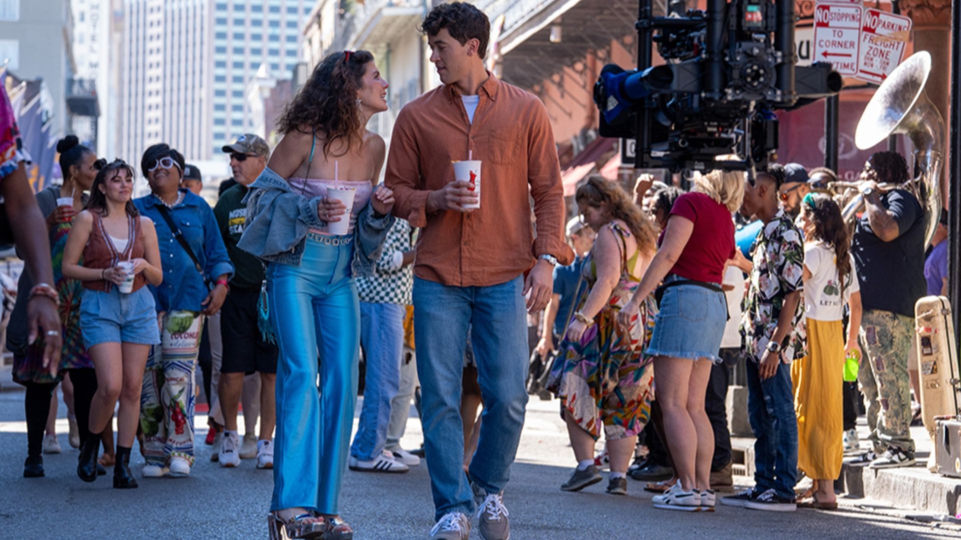 A young couple walking on a city street holding drinks, smiling at each other, surrounded by a crowd with colorful clothing, and a camera rig overhead in a lively urban setting.