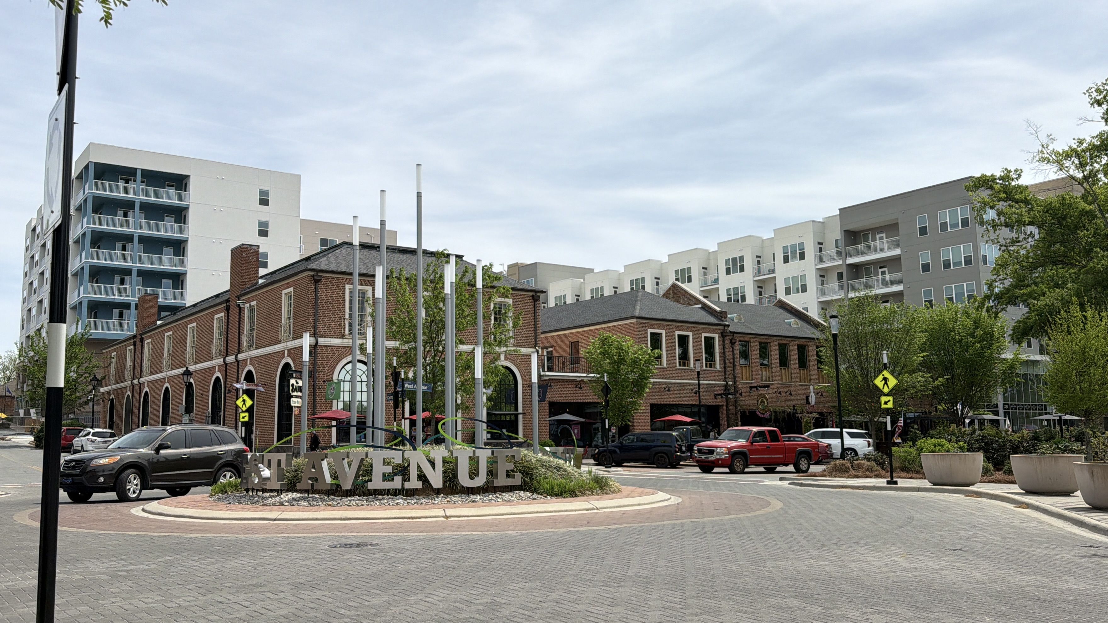 Brick shopping block around a roundabout with a large AVENUE sign in the foreground. Behind it are modern white and gray apartment buildings, trees, and parked cars under a blue sky.