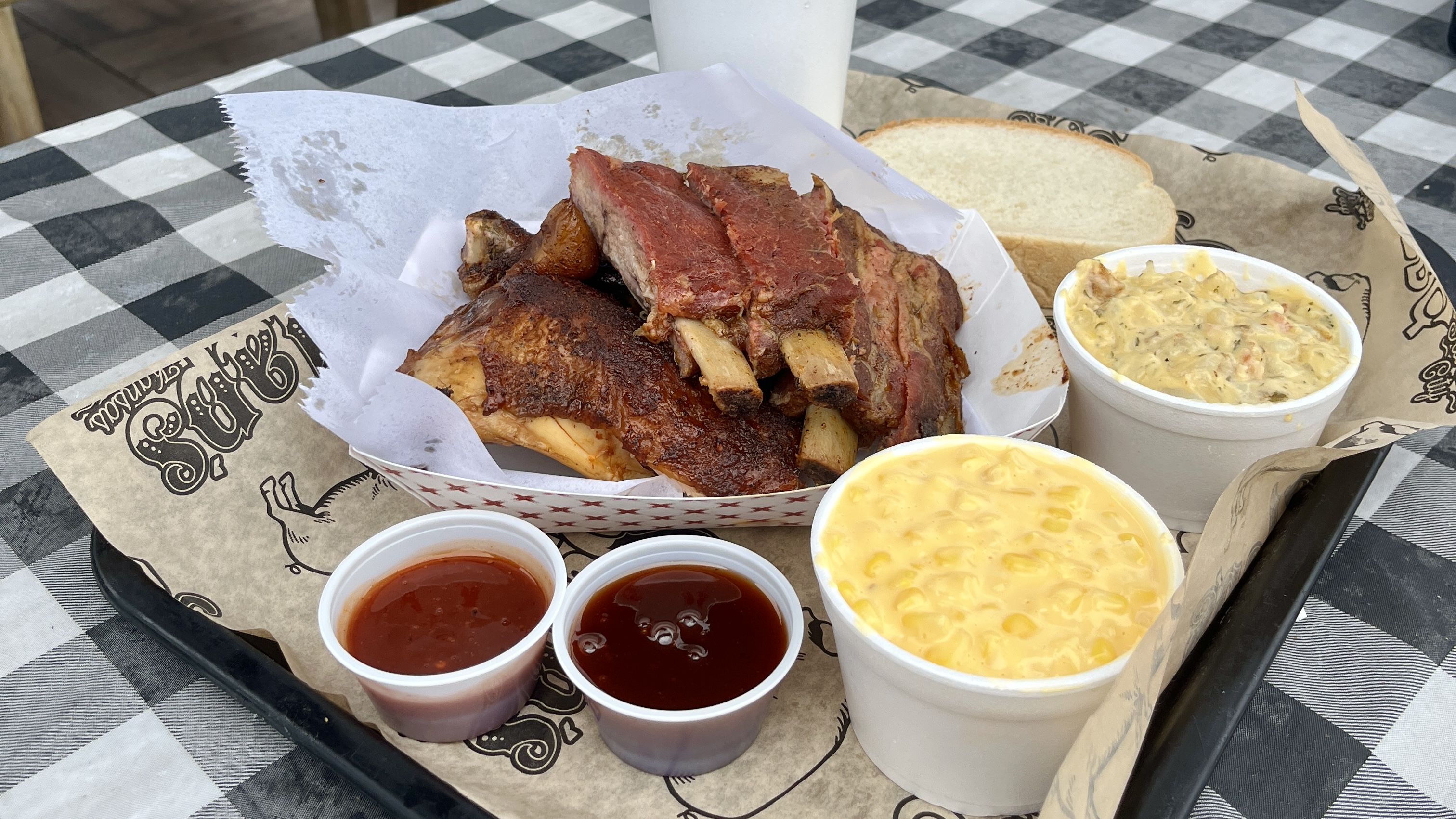 A tray of food includes ribs, chicken, two sauces, cheesy corn, baked potato casserole, and white bread on a checkered tablecloth.