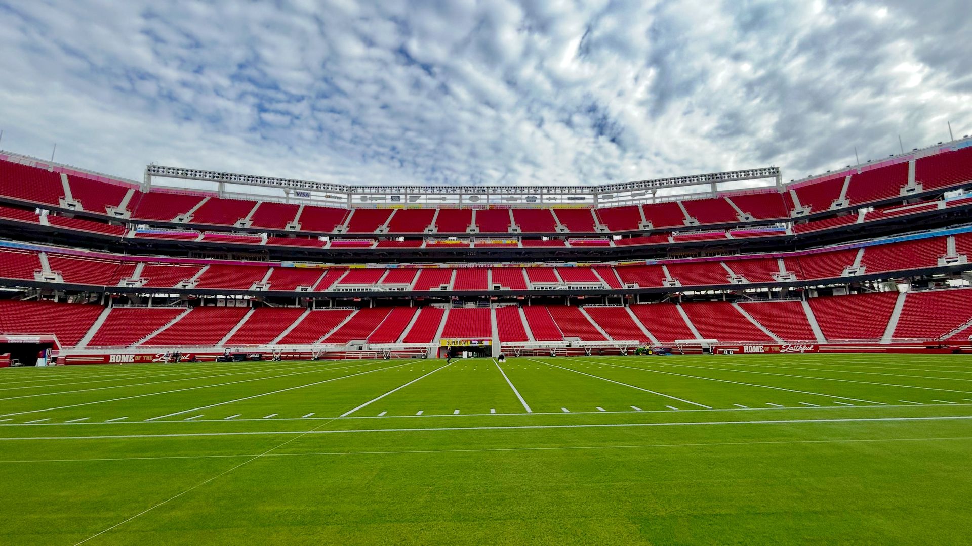 Empty football stadium with bright red seats, green field marked with white lines, under a cloudy blue sky. Signs say "HOME OF THE Faithful" and "SUPER BOWL."