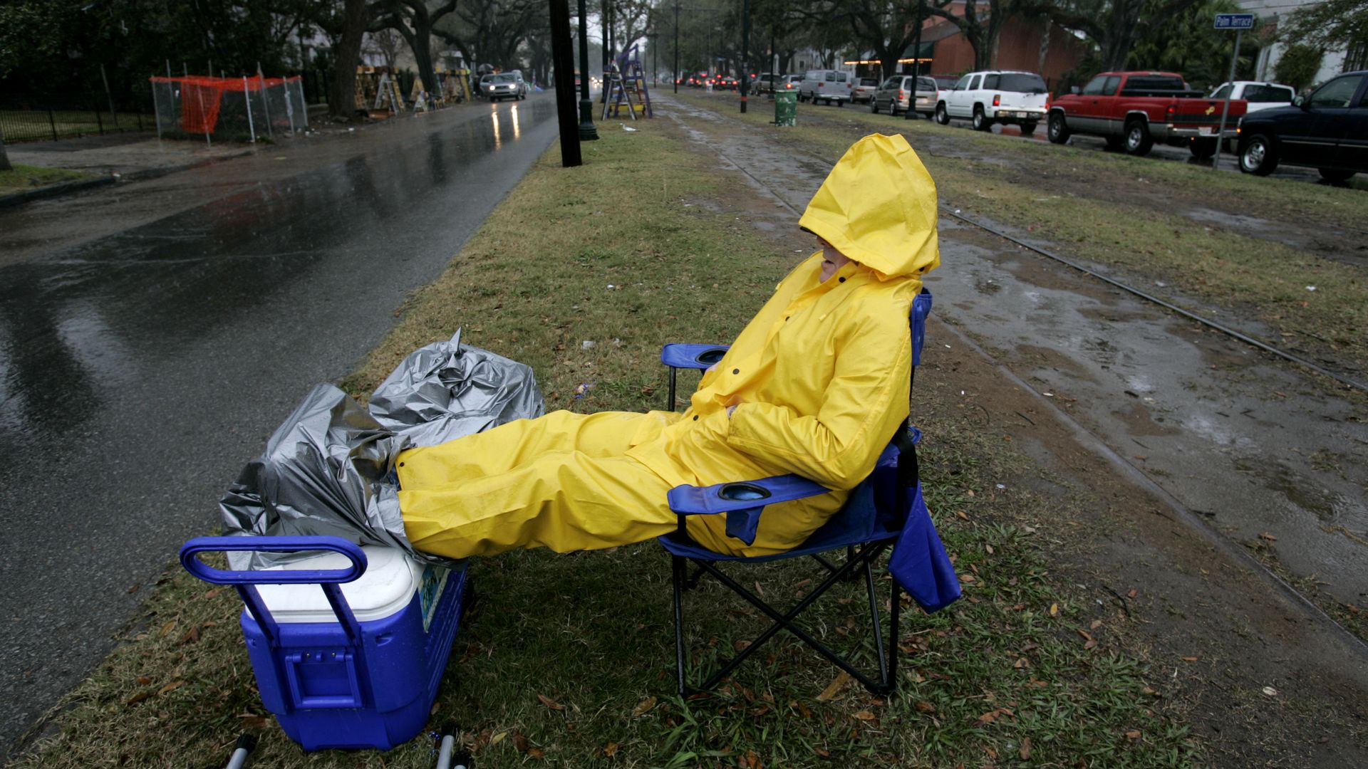 A would-be parade-goer wears a rain jacket and pants as they sit, huddled up, in the rain on the neutral ground.