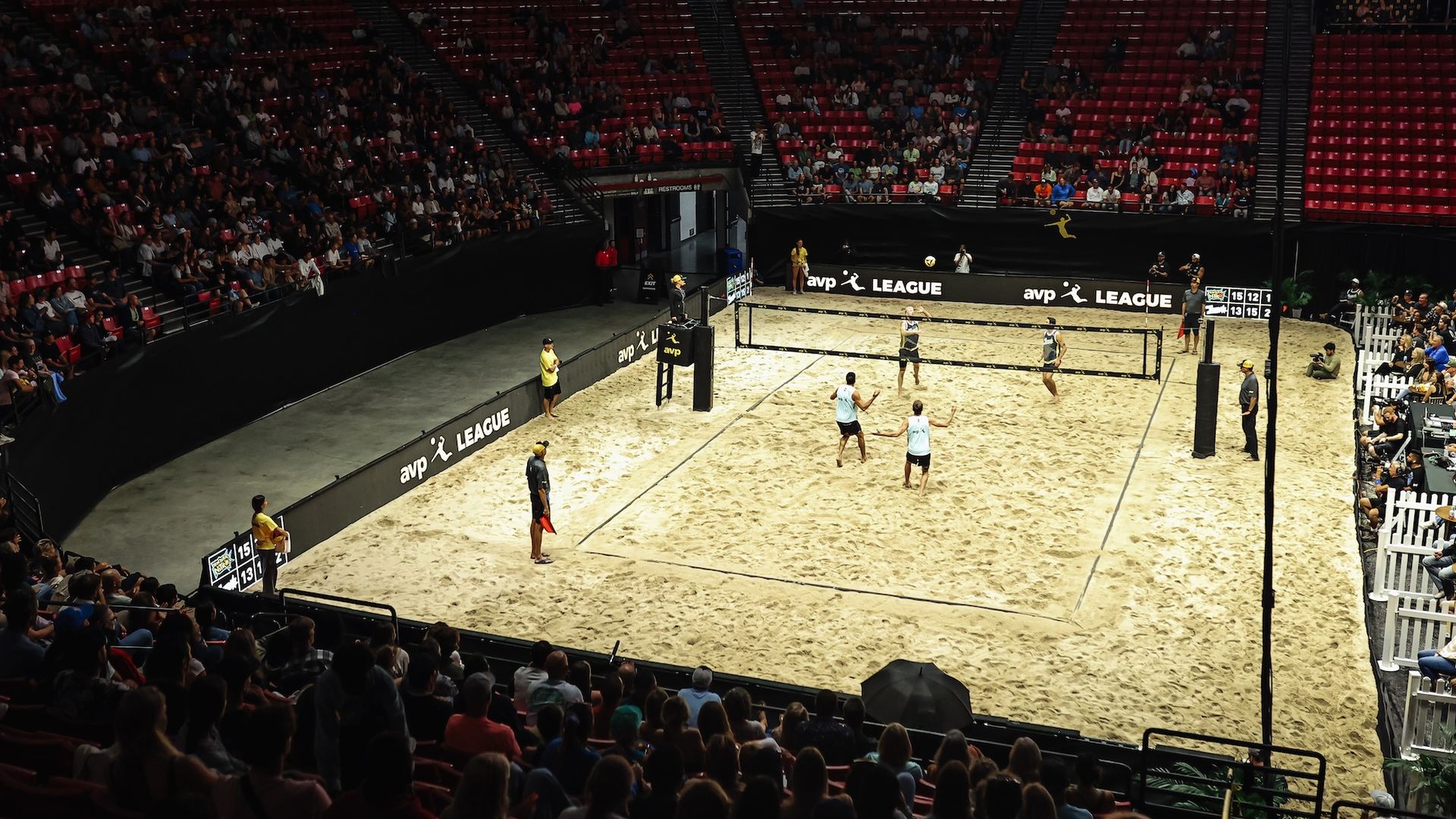 A beach volleyball court is set up for a match inside an arena with fans in the stands and players on the sand.