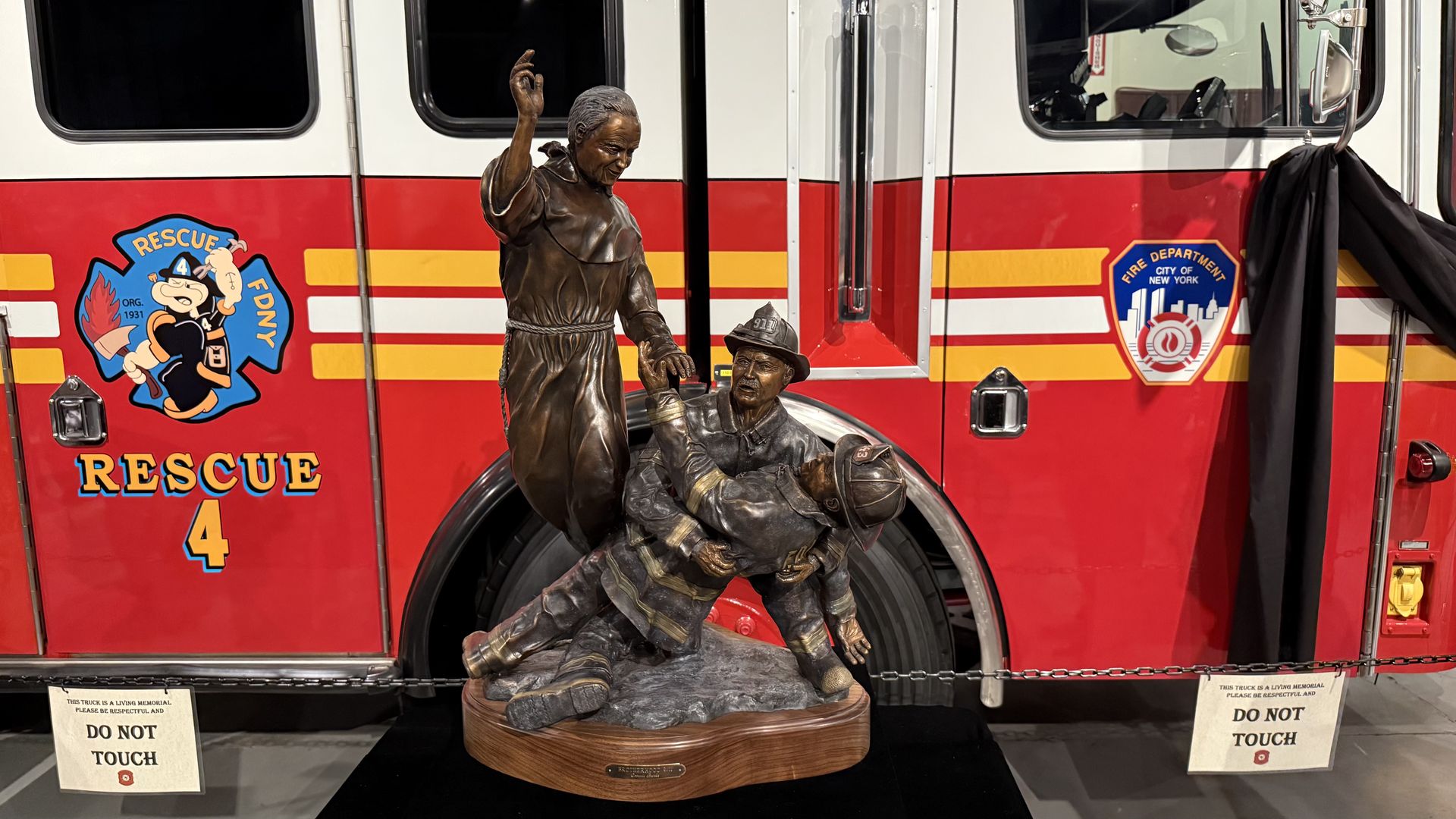 Bronze statue of a priest standing over two firefighters, one of whom is holding the other. The statue is on a table in front of a fire truck.