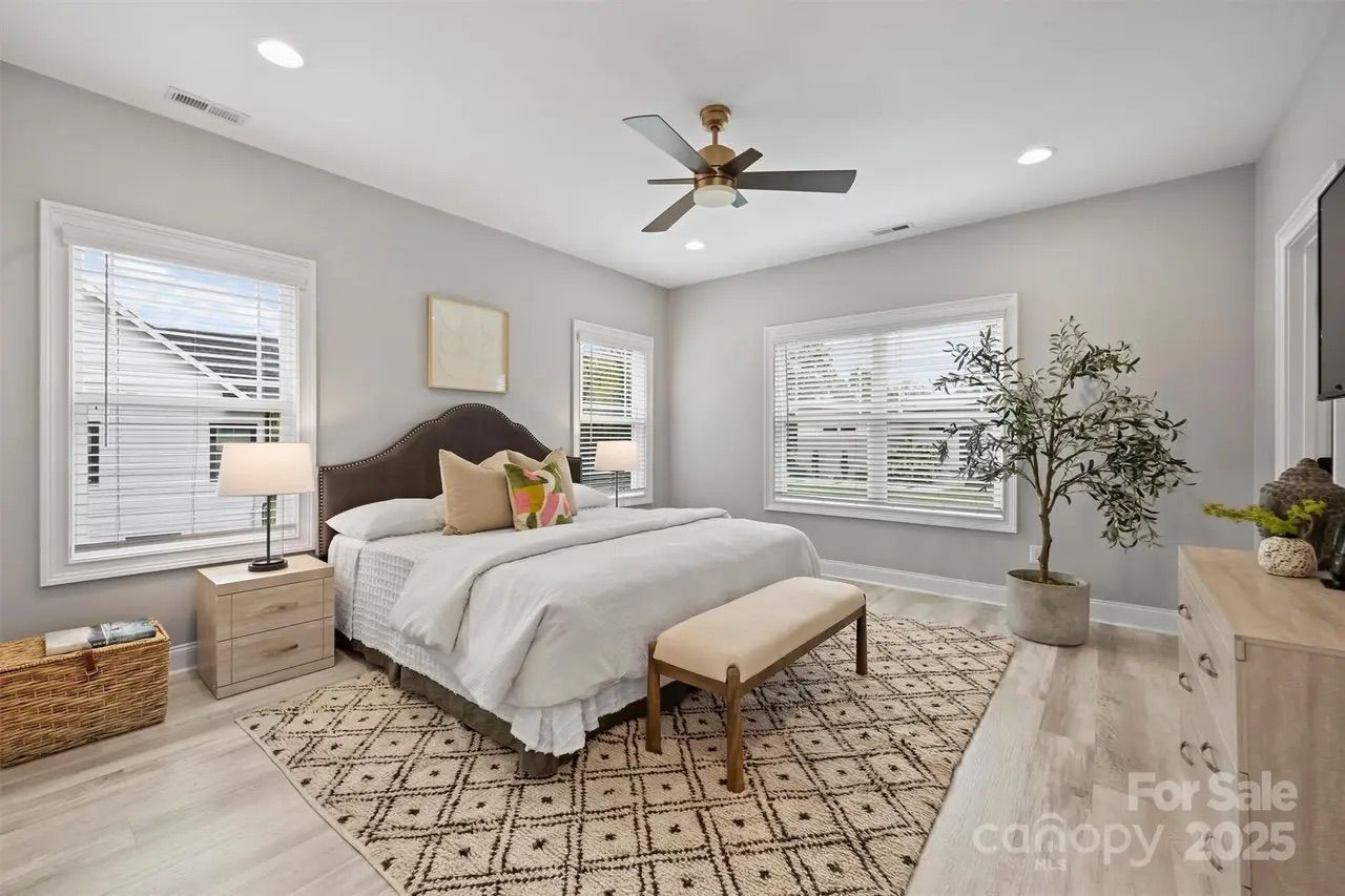 Bright bedroom with gray walls, large bed with white bedding and colorful pillows, beige bench, patterned rug, two nightstands with lamps, ceiling fan, large windows, and potted plant.