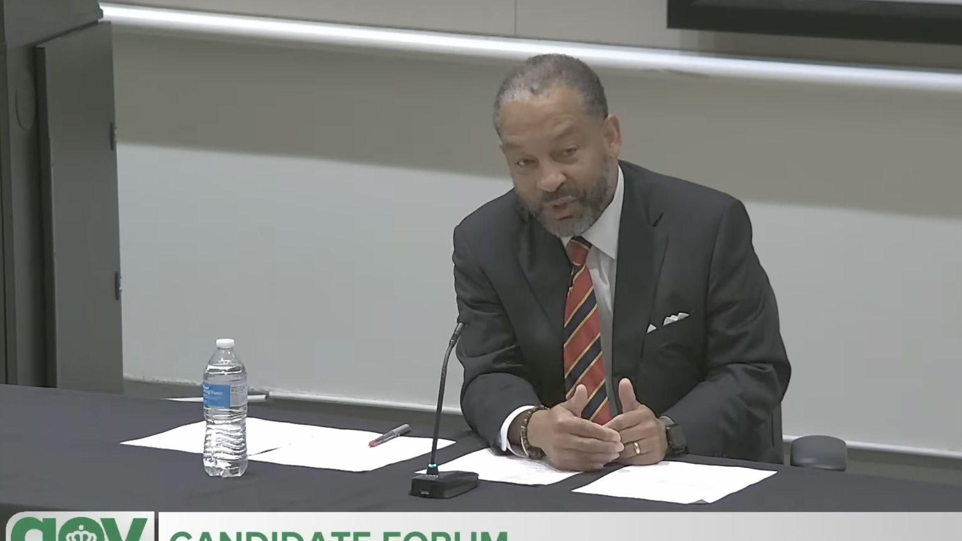 Bearded man in a dark suit and striped tie sits at a table with papers and a microphone during a city council candidate forum; a water bottle and a banner reading "CANDIDATE FORUM" are visible.