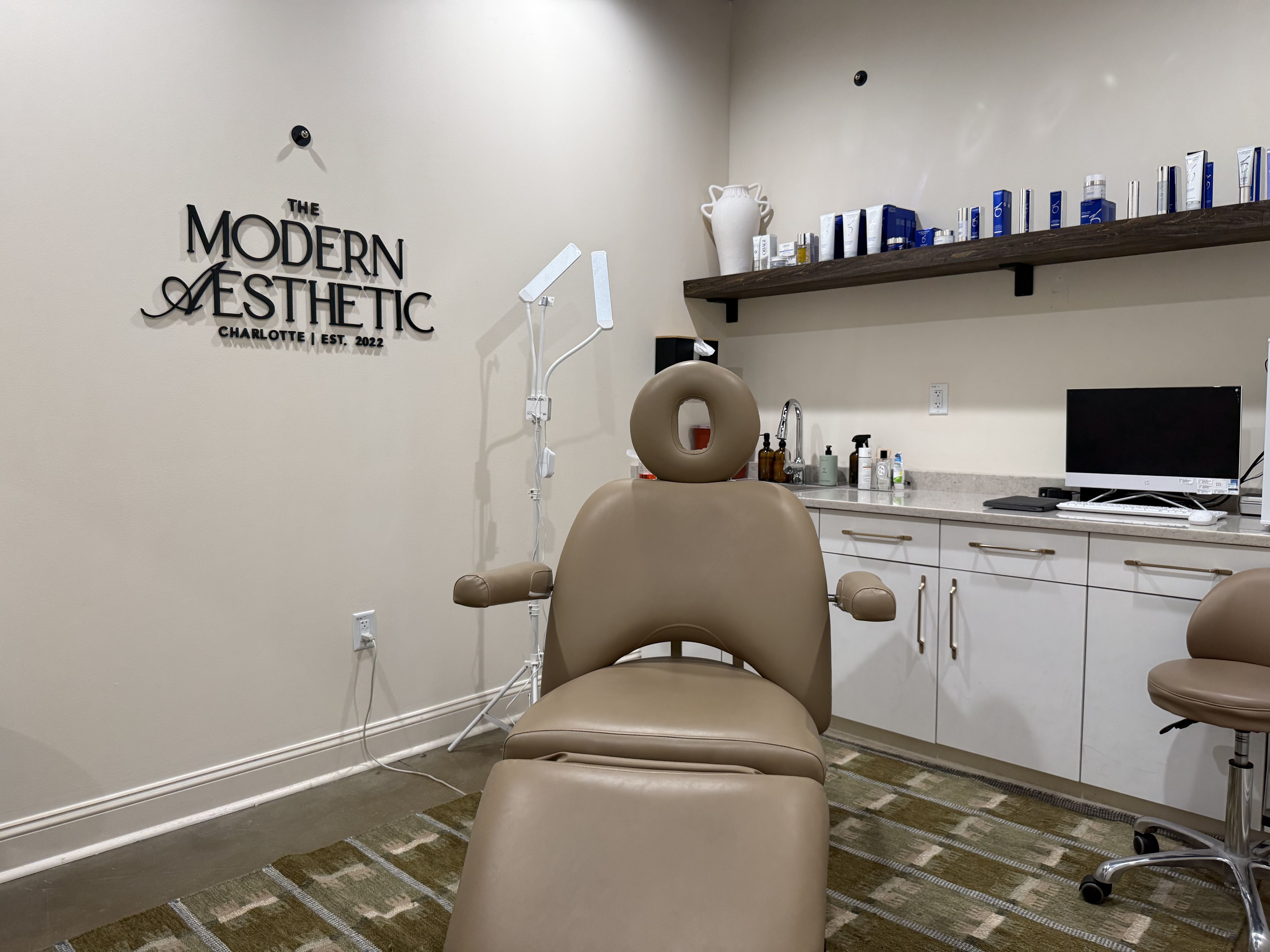 Interior of a modern aesthetic clinic with beige walls, a sign reading The Modern Aesthetic, beige treatment chairs, and a long wooden shelf of blue tubes above a counter with a computer.