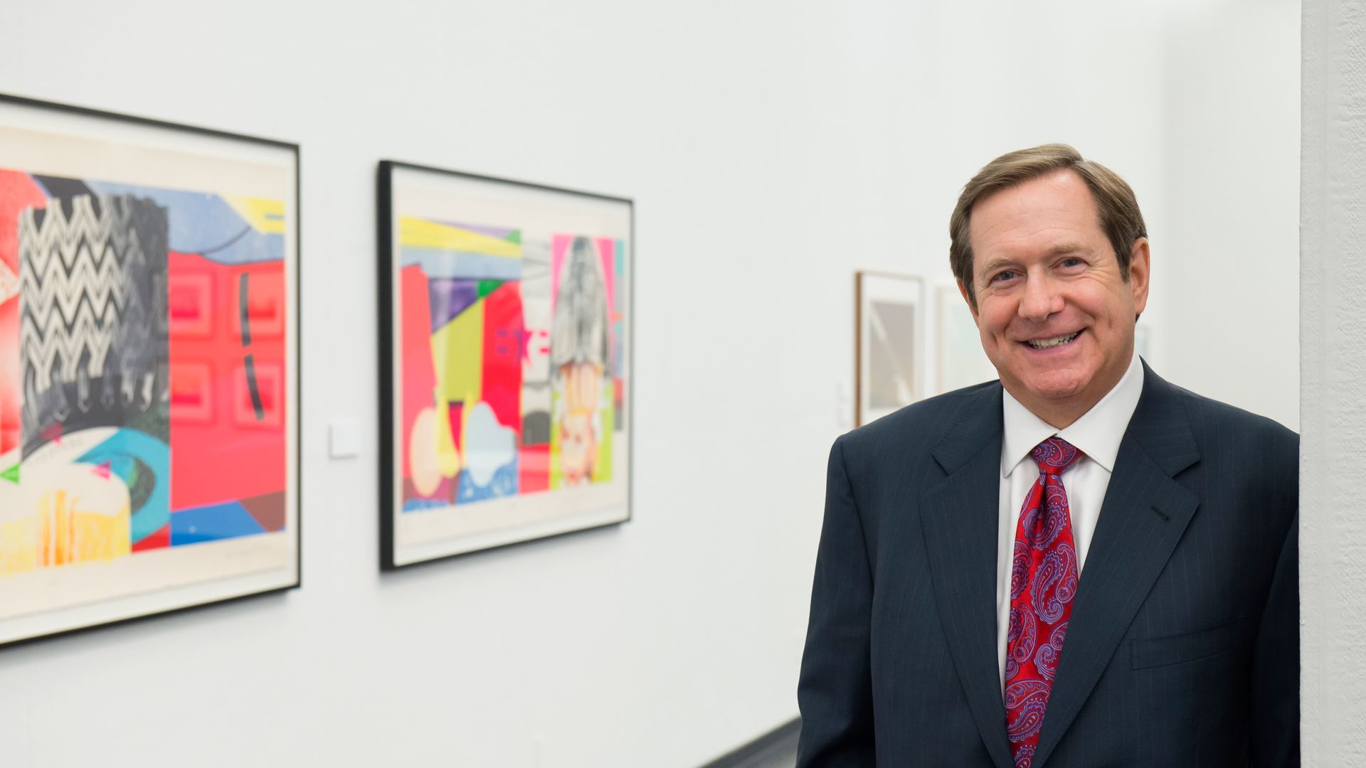 Middle aged white man in suit and tie standing in art gallery in front of three framed prints