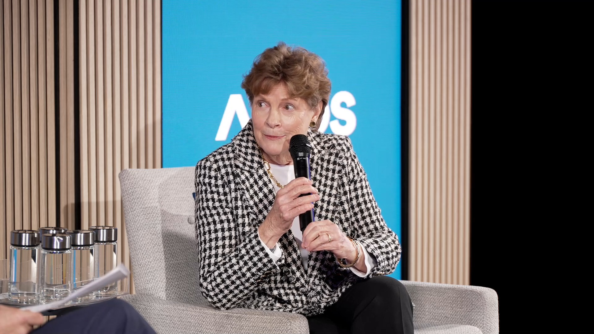 Woman with short curly brown hair wearing a black-and-white houndstooth jacket speaking into a microphone while sitting on a gray upholstered chair against a blue background.