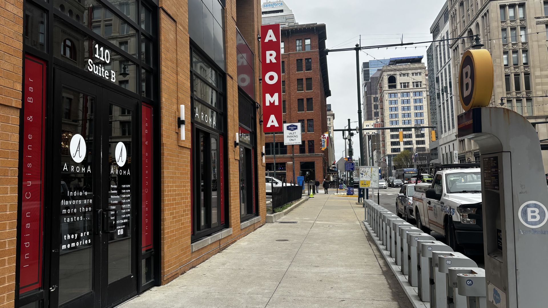 City street with brick storefronts, a tall red AROMA sign, glass doors with circular logos, and 110 Suite B on the windows. A blue-and-yellow B sign tops a transit kiosk as cars line the curb.